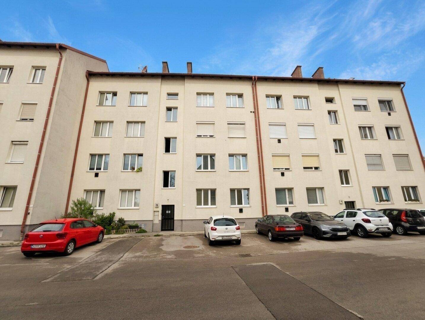 Mehrfamilienhaus mit heller Fassade und Parkplätzen vor dem Gebäude.
