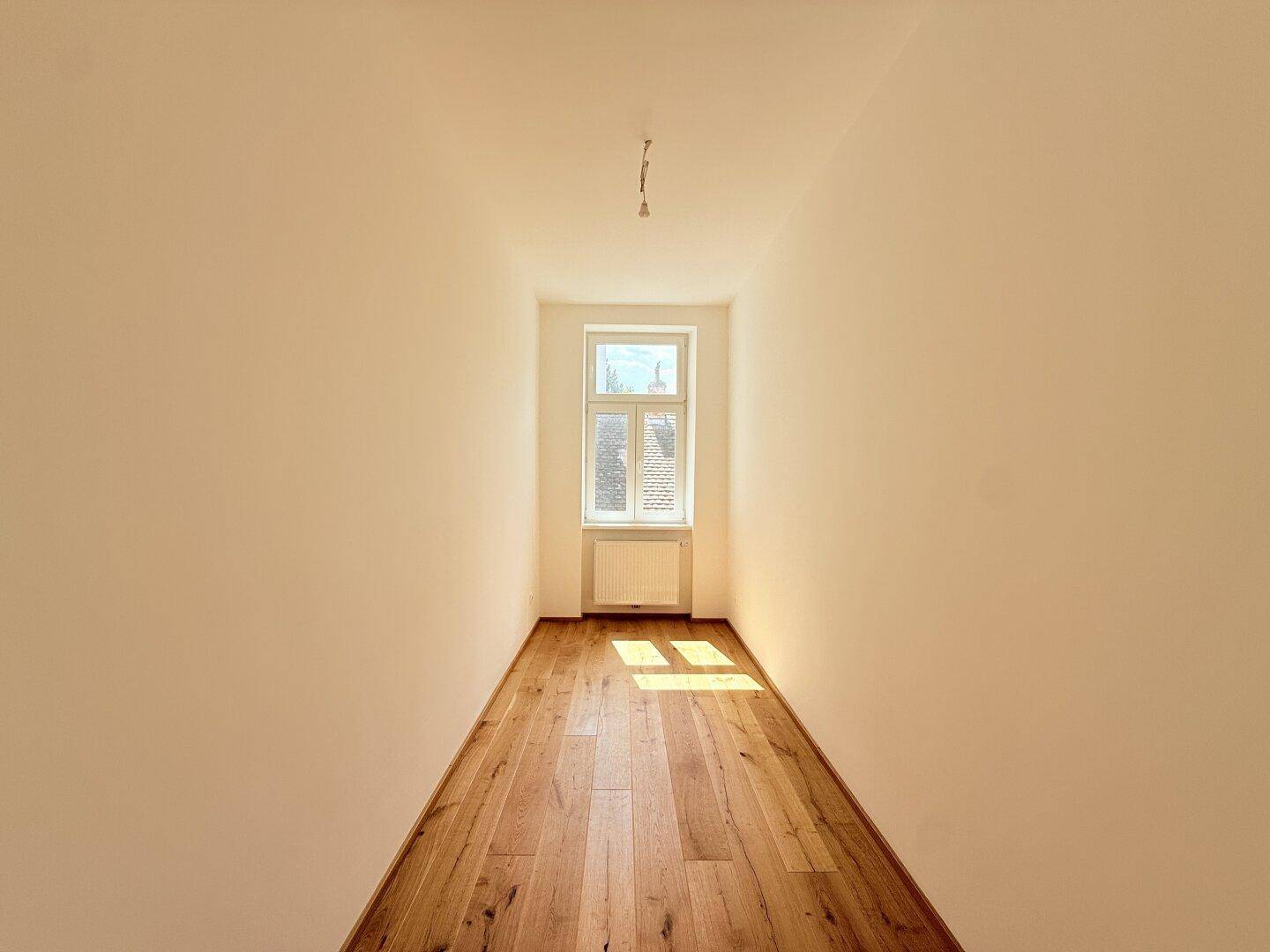 Schmaler, heller Raum mit Holzboden und einem Fenster am Ende, bietet Potenzial für verschiedene Nutzungen.