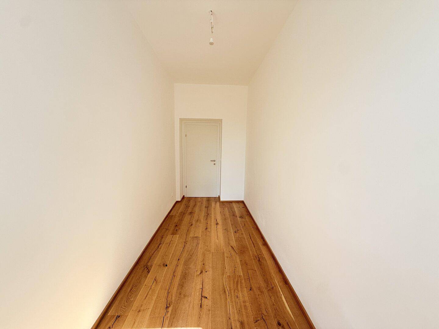 Heller Flur mit Holzboden und weiß gestrichenen Wänden, führt zu einer weiteren Tür im Hintergrund.