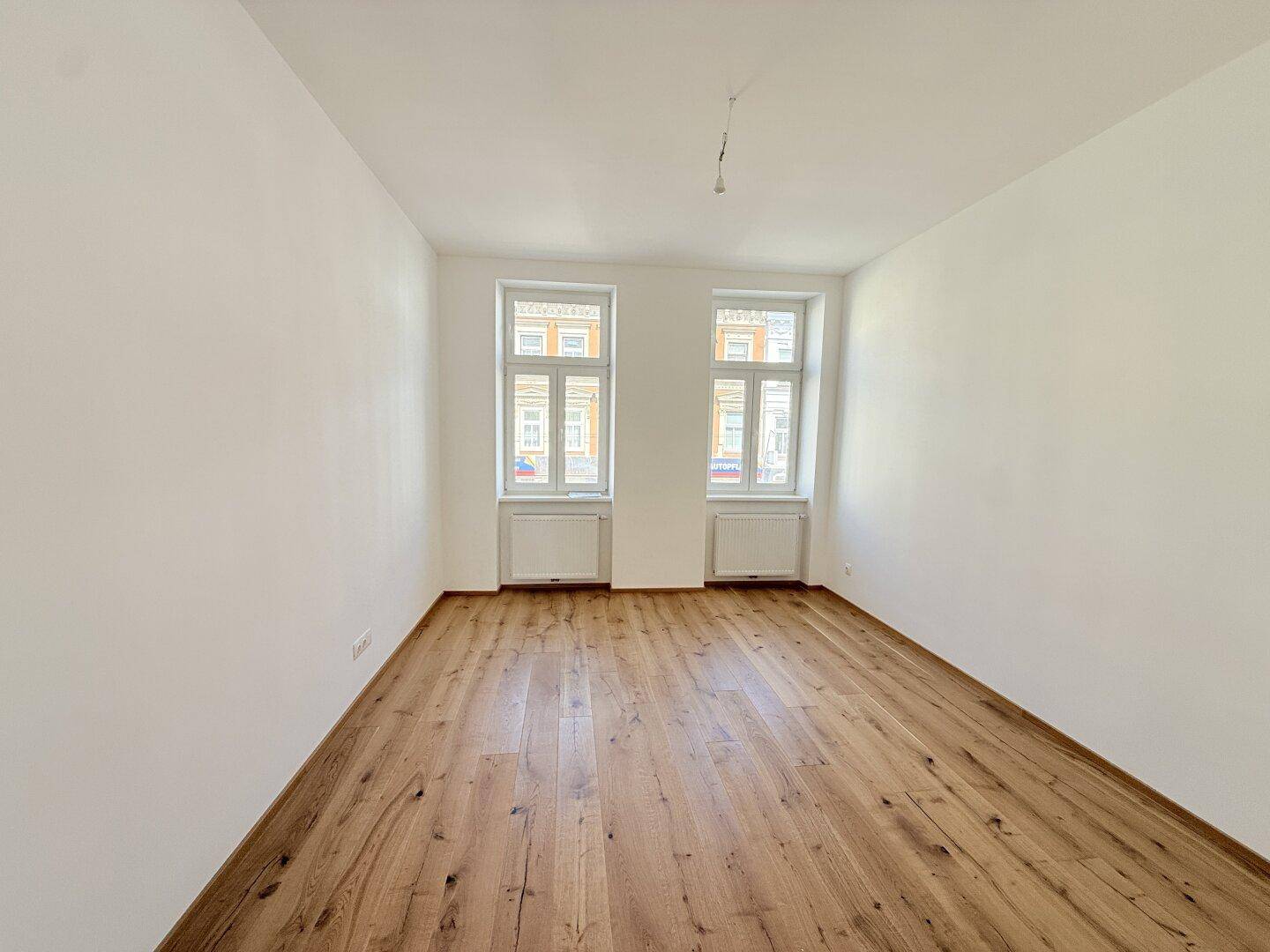 Geräumiger, heller Raum mit Holzboden und zwei Fenstern, ideal für Wohnzwecke.