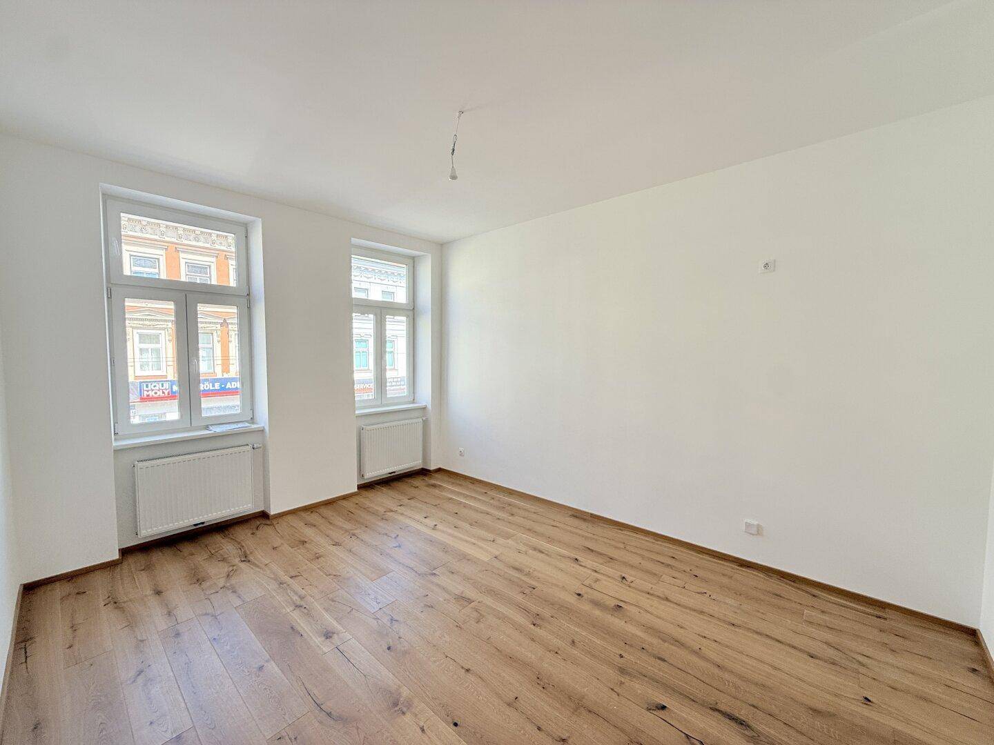 Heller, leerer Raum mit Holzboden und zwei Fenstern, bietet viel Tageslicht.