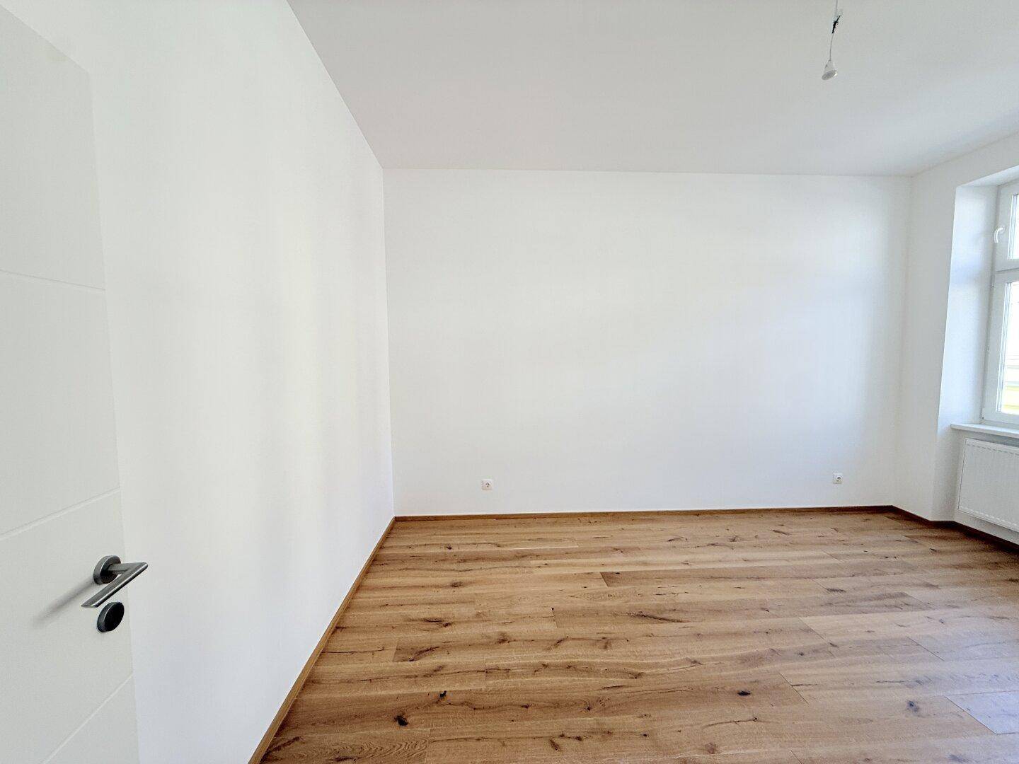 Leerer Raum mit Holzboden, weiß gestrichenen Wänden und einem Fenster.