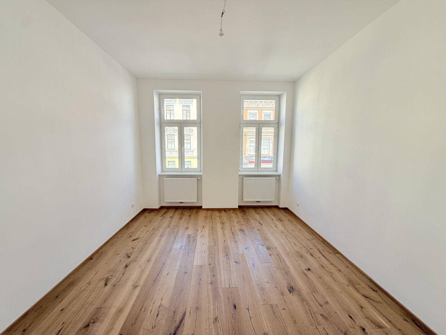Geräumiger, heller Raum mit Holzboden und zwei Fenstern, ideal für Wohnzwecke.