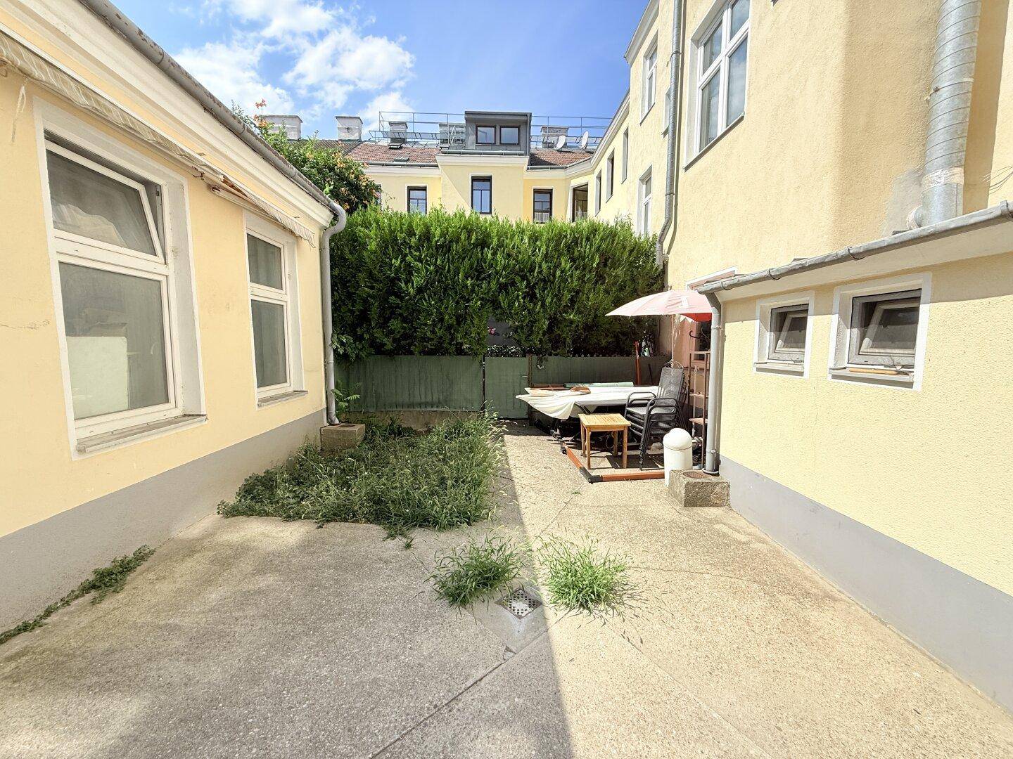 Hinterhof mit gepflastertem Bereich, Grünflächen und einer kleinen Sitzecke unter einem Sonnenschirm.