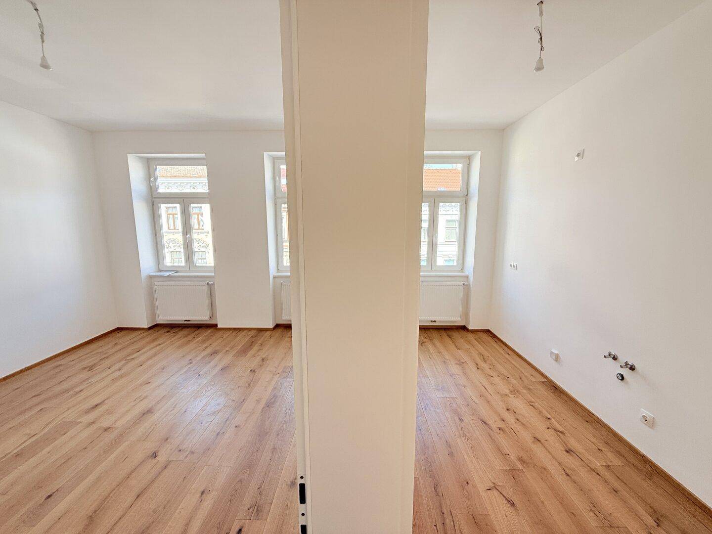Heller Raum mit Holzboden und Säule, vorbereitet für Kücheninstallationen, mit zwei Fenstern.