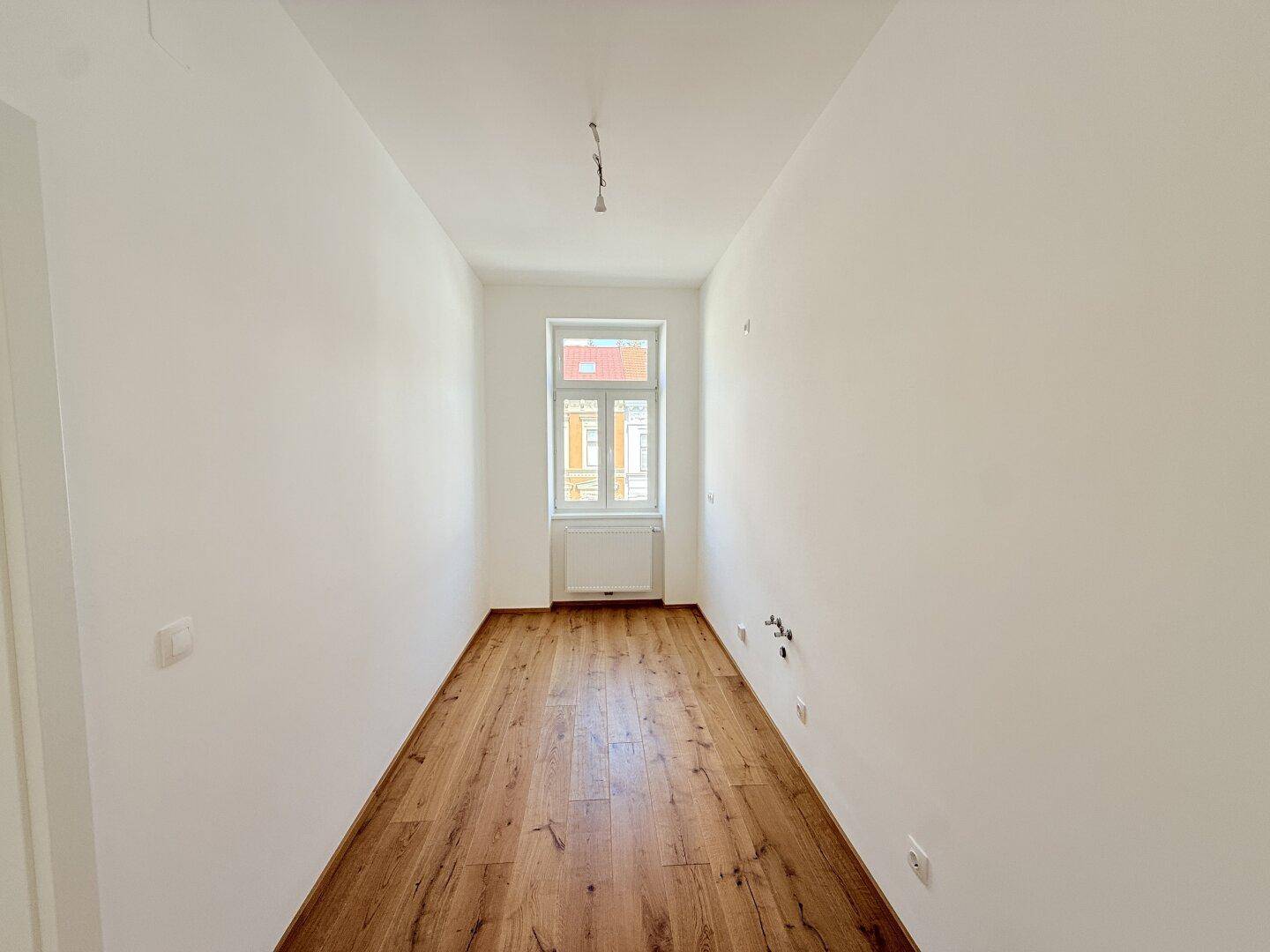 Schmaler, heller Raum mit Holzboden und einem Fenster, vielseitig nutzbar als kleines Büro.