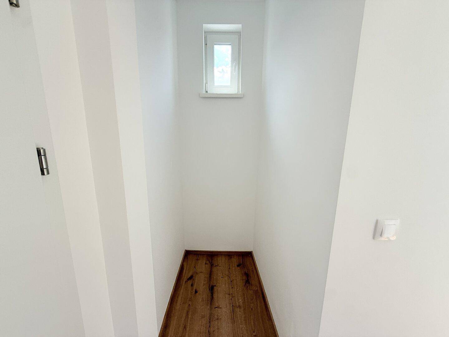 Schmaler Raum mit kleinem Fenster und Holzboden, ideal als Abstellraum oder Speisekammer.