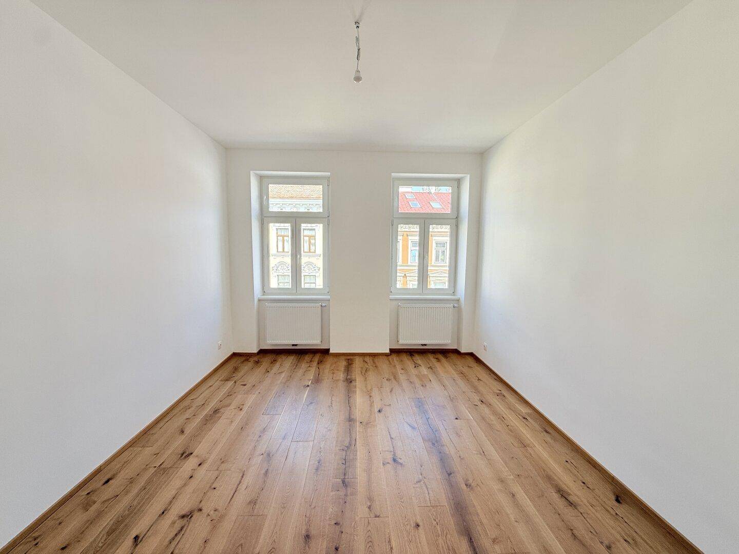 Heller, leerer Raum mit Holzboden und zwei Fenstern, ideal für ein großzügiges Wohnzimmer.