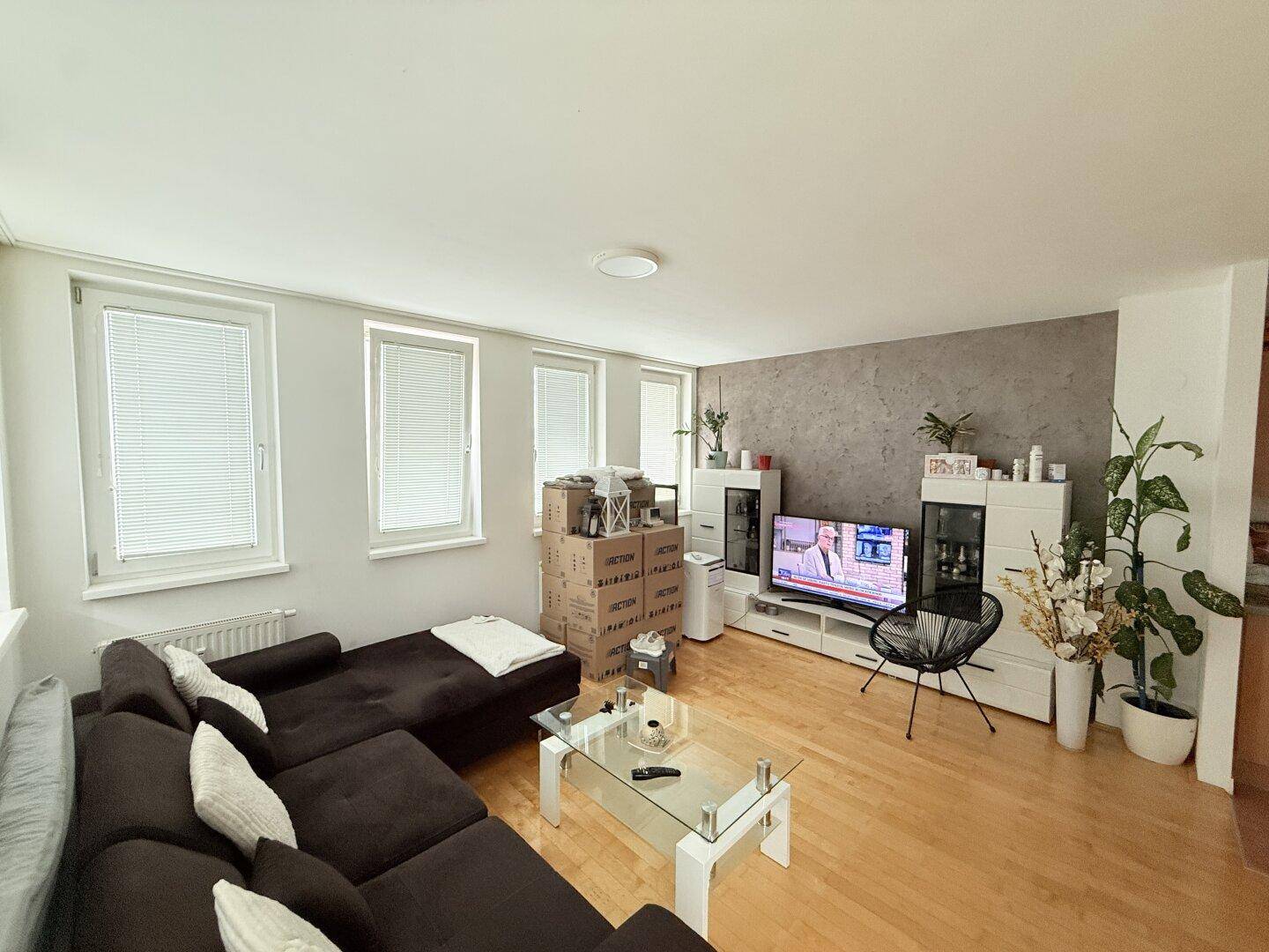 Gemütliches Wohnzimmer mit dunklem Ecksofa, Holzboden und vielen Fenstern.