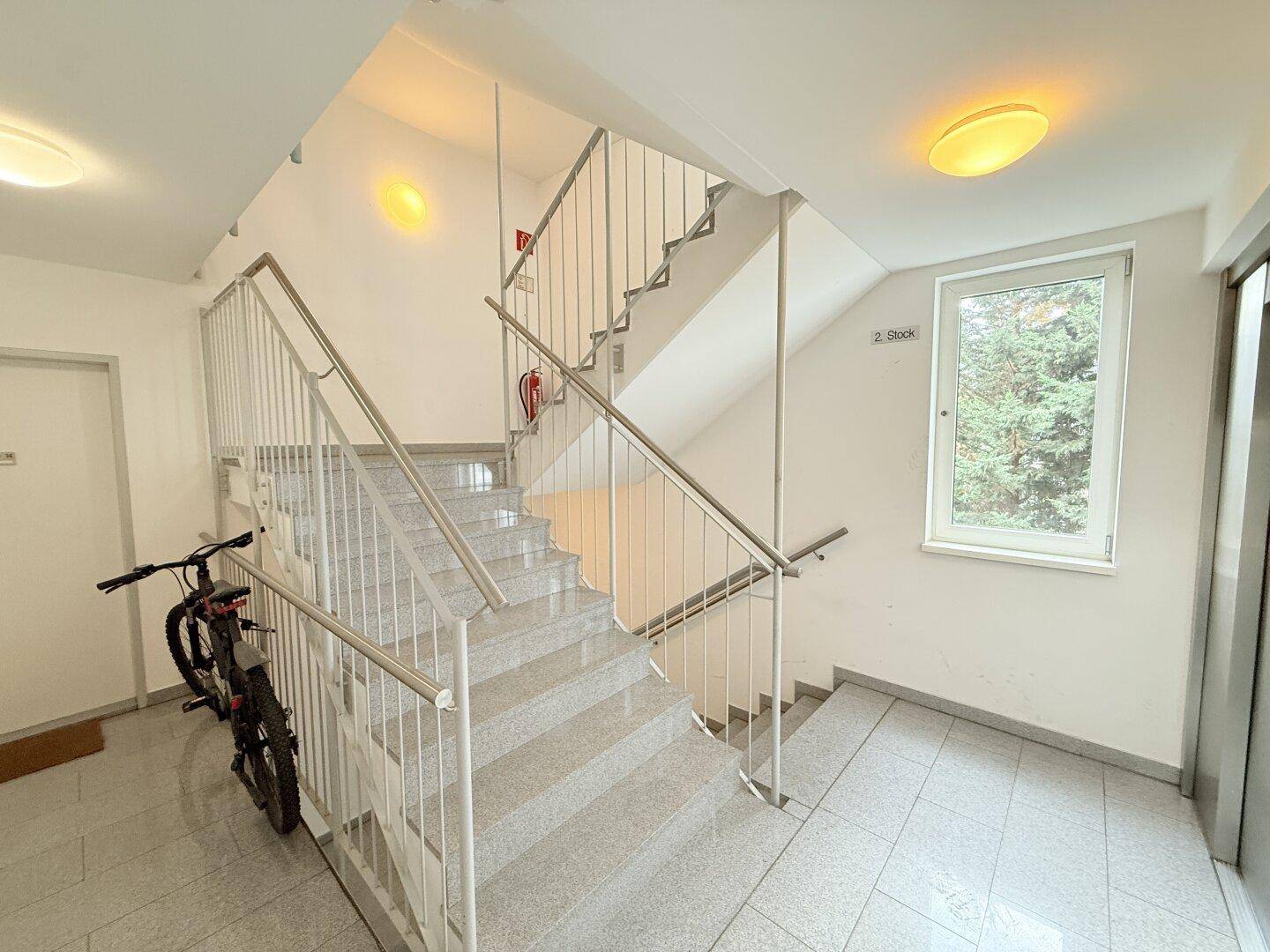 Treppenhaus mit Granitstufen, Metallgeländer und Fenster, Fahrrad abgestellt.