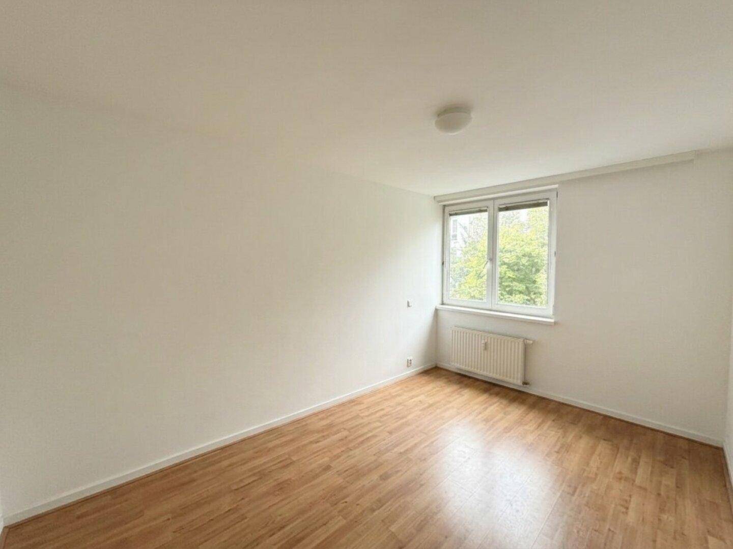 Helles Schlafzimmer mit Holzboden, Fenster und Heizkörper, Blick ins Grüne.