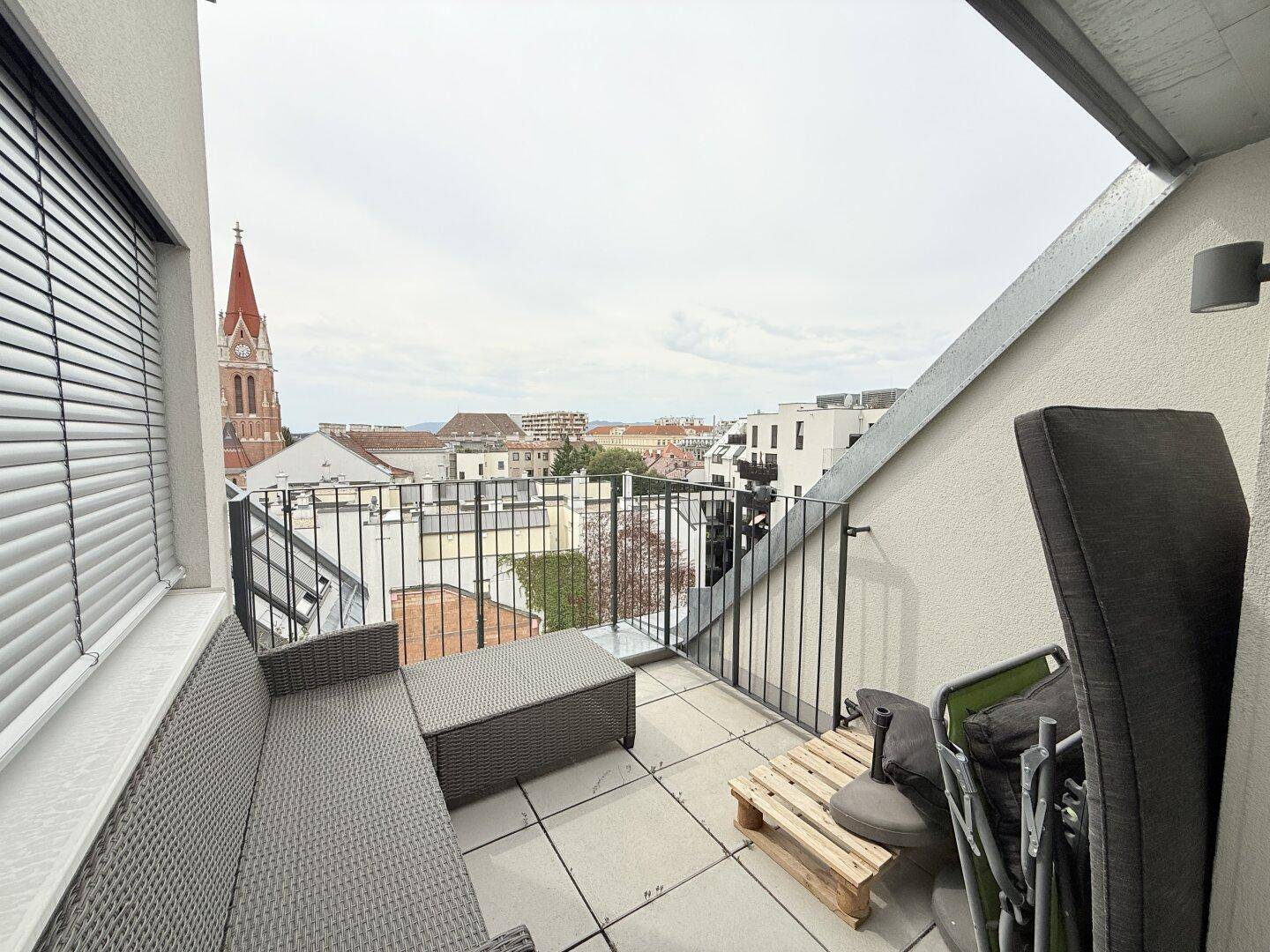 Geräumiger Balkon mit Sitzgelegenheiten und weitem Blick über die Stadt und einen Kirchturm.