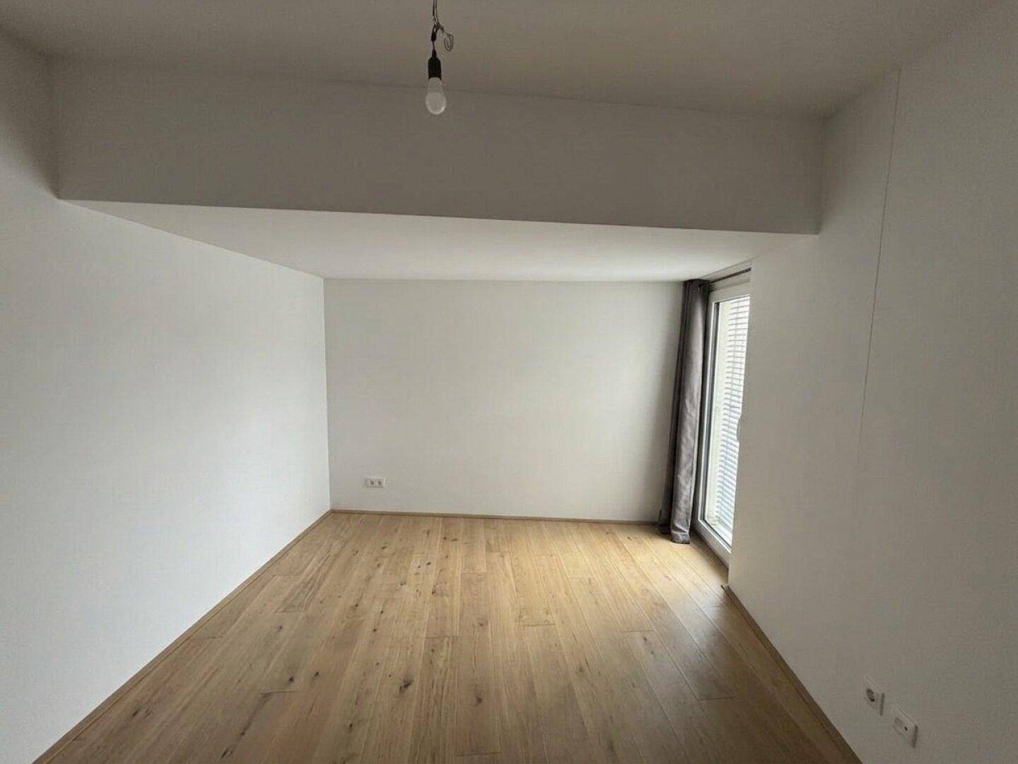 Helles, leeres Zimmer mit Holzboden und Fenster mit Jalousien.