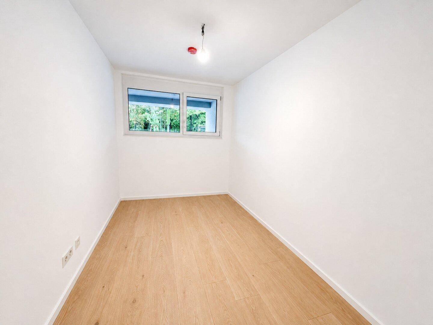Leeres Schlafzimmer mit hellem Holzboden und Fenster, das einen grünen Ausblick bietet.