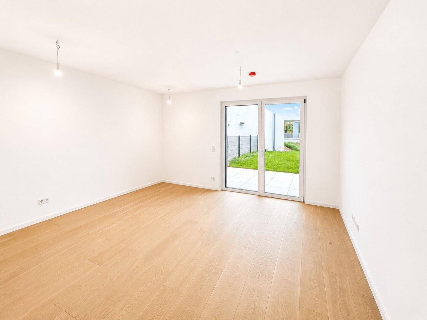 Heller, leerer Wohnraum mit Holzfußboden und großen Fenstern zum Garten.