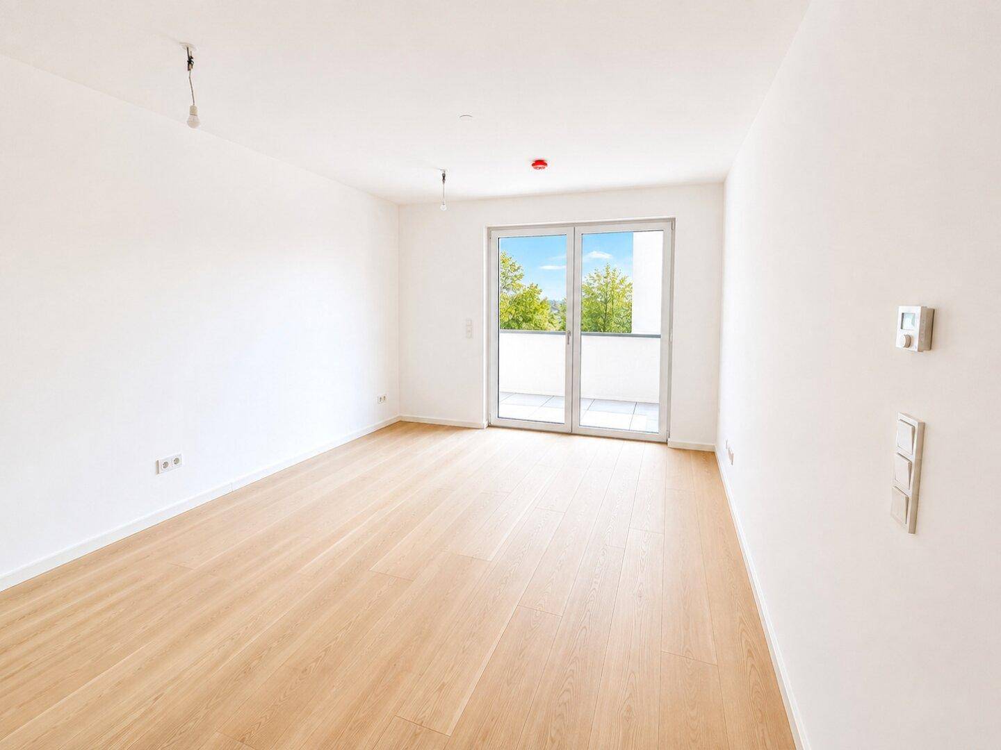 Helles, leeres Wohnzimmer mit hellem Holzparkett und Zugang zu einem Balkon.