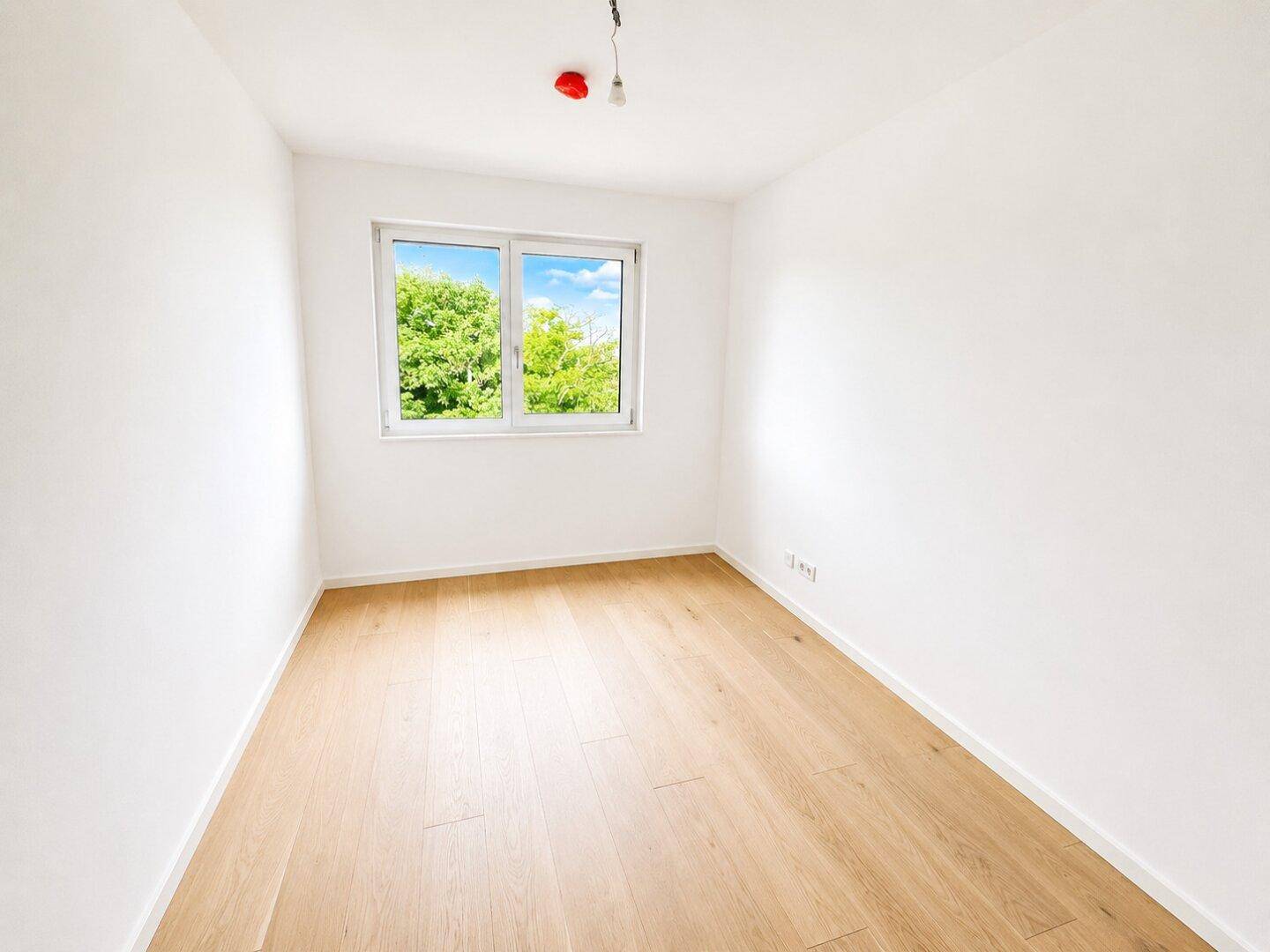 Unmöbliertes Zimmer mit hellem Holzparkett und großem Fenster, das viel Tageslicht hereinlässt.