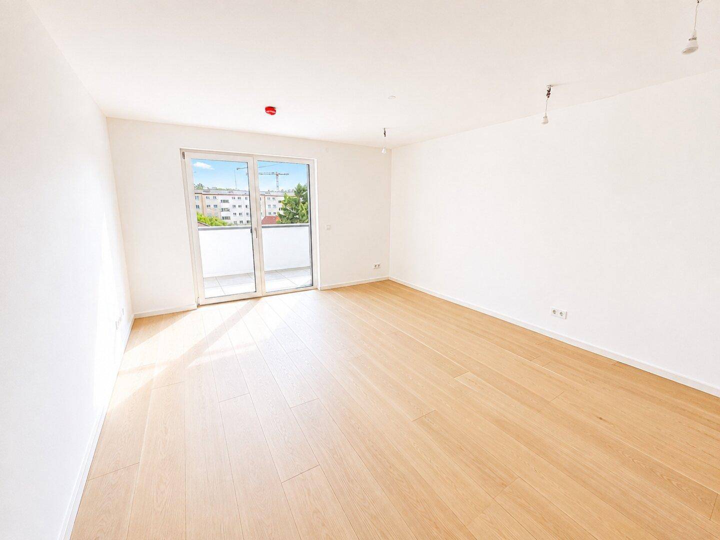 Helles, leeres Zimmer mit hellem Holzparkett und Zugang zu einem Balkon.