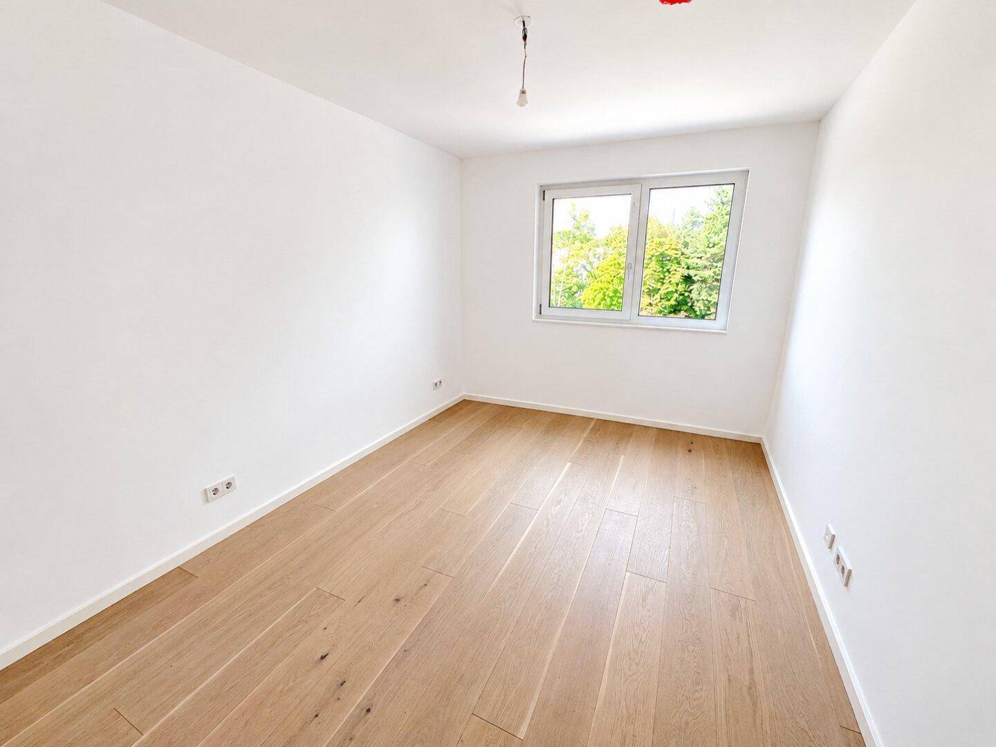 Ruhiges, leeres Zimmer mit hellem Holzparkett und Fenster mit Blick ins Grüne.