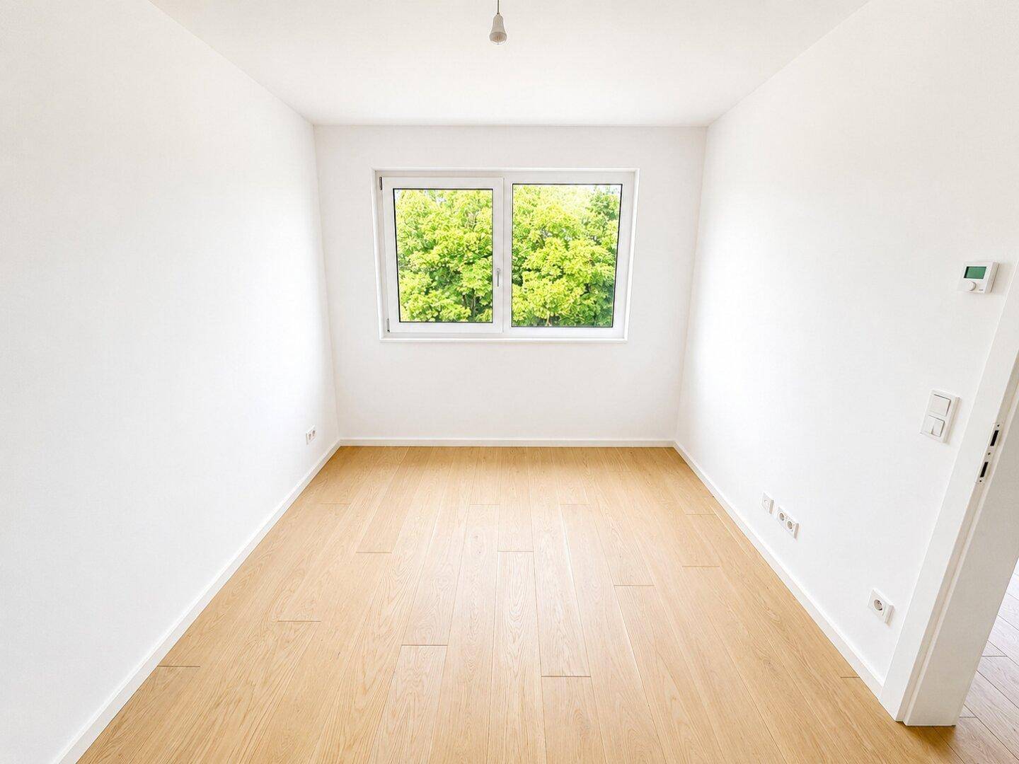 Helles, leeres Zimmer mit weiß gestrichenen Wänden, Holzparkett und Fenster mit Blick ins Grüne.