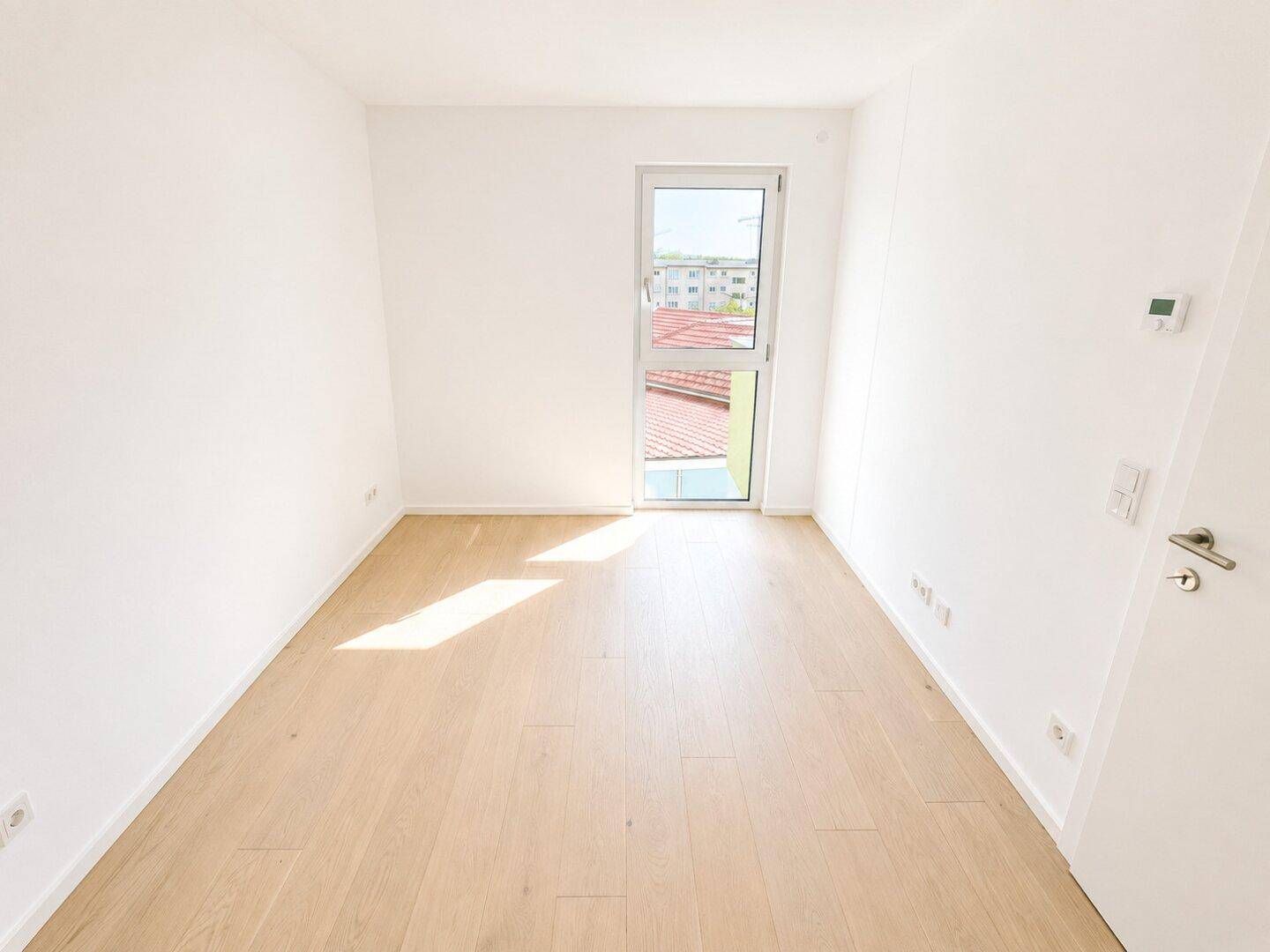 Helles, leeres Zimmer mit Parkettboden und einem Fenster, das Tageslicht hereinlässt.