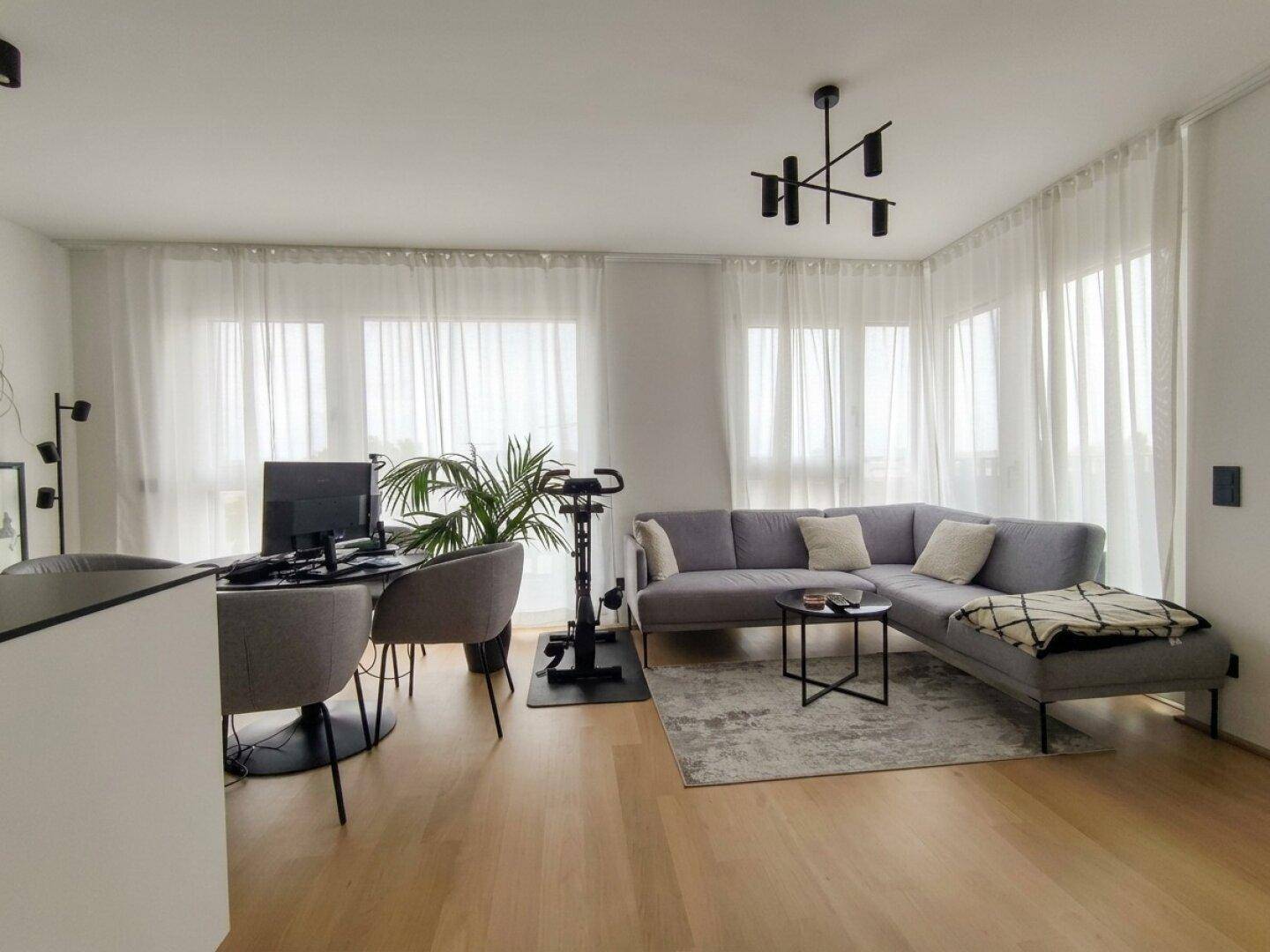 Geräumiger Wohnbereich mit bequemer Couch, Arbeitsplatz und viel Tageslicht durch die großen Fenster.