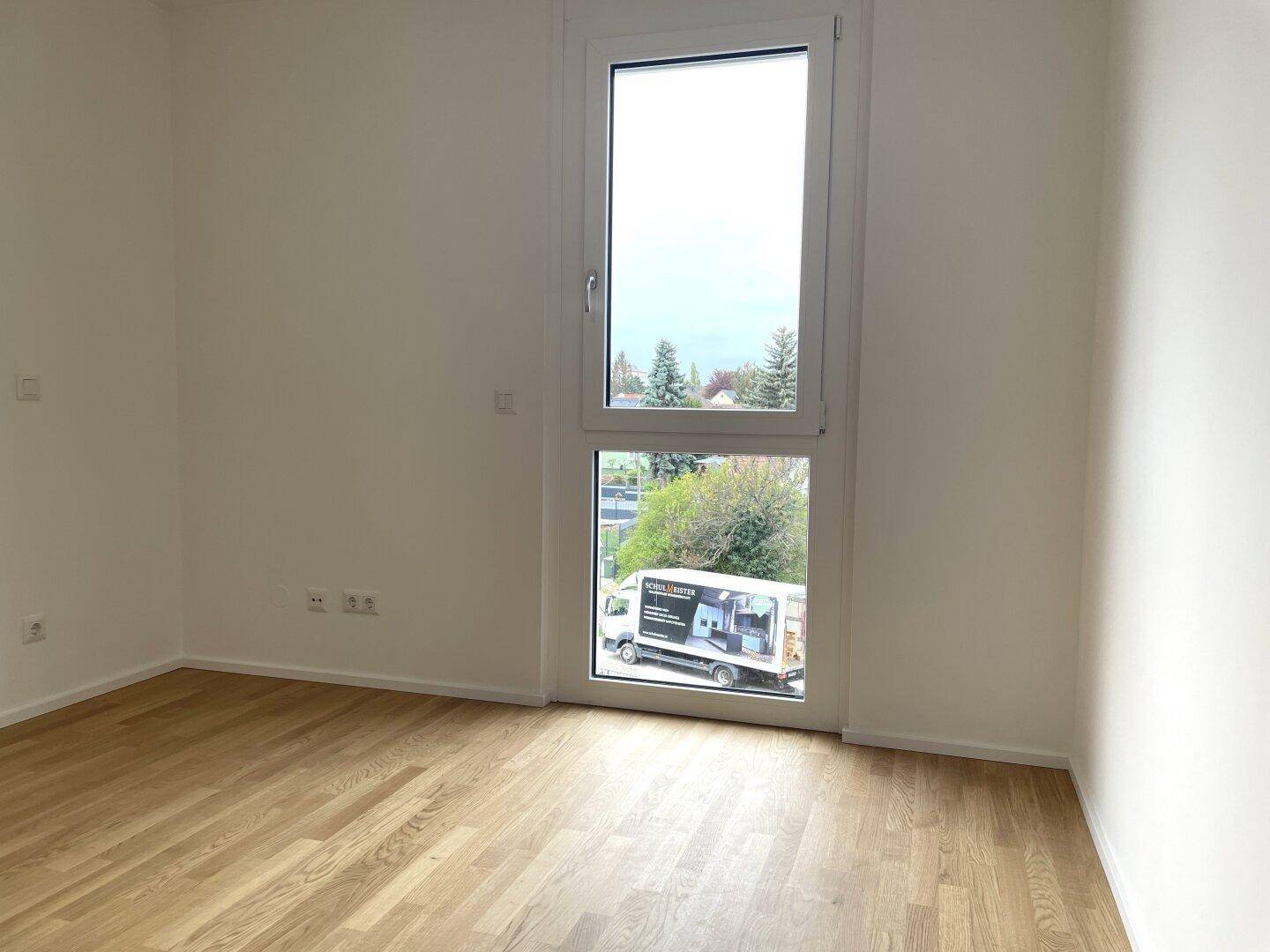 Heller Raum mit Holzboden und hohem Fenster, das einen Blick auf die Umgebung ermöglicht.