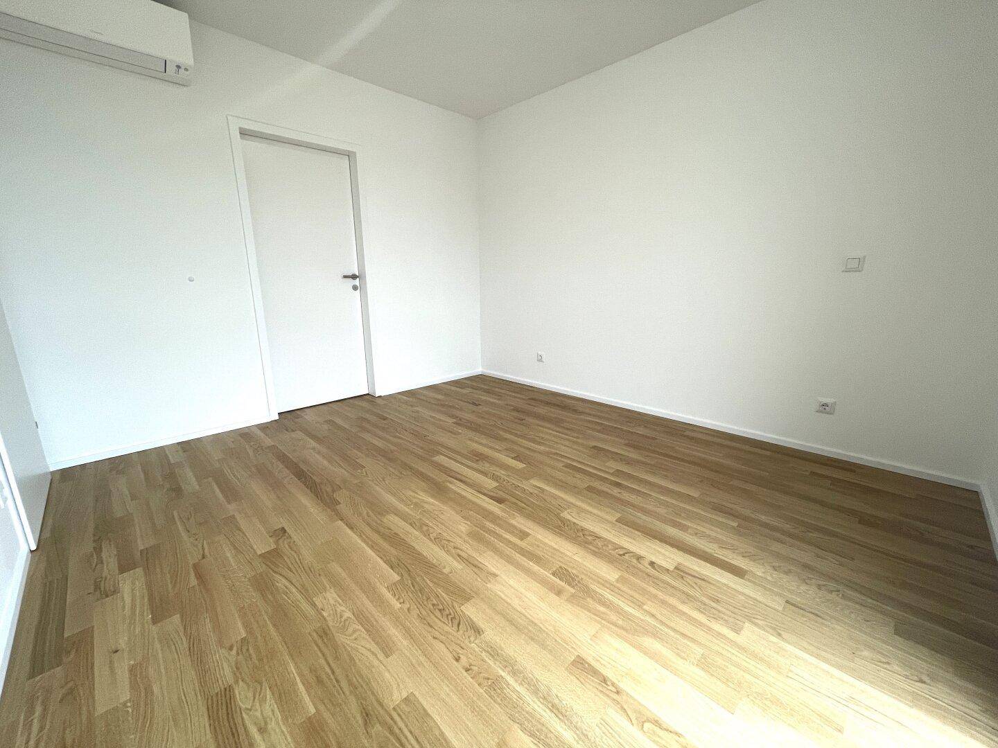 Schlichter, leerer Raum mit Holzboden und weißer Tür, ideal als Schlaf- oder Arbeitszimmer.