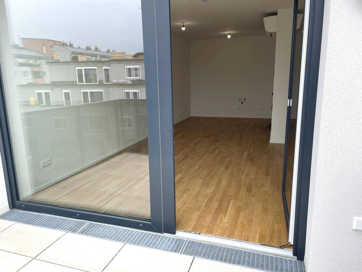 Blick von der Terrasse in einen hellen Innenraum mit Holzboden durch eine große Schiebetür.