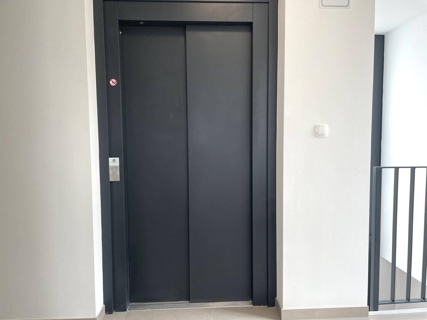 Moderner Aufzug mit dunklen Türen in einem hellen Treppenhaus, bietet bequemen Zugang zu allen Etagen.