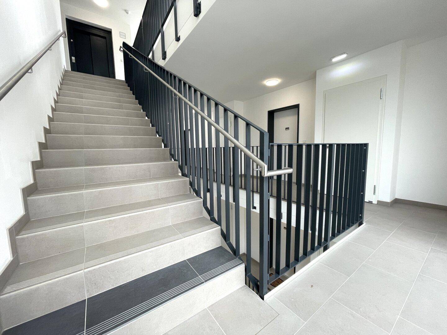 Helles Treppenhaus mit modernen Fliesenstufen und dunklem Metallgeländer in einem Neubau.