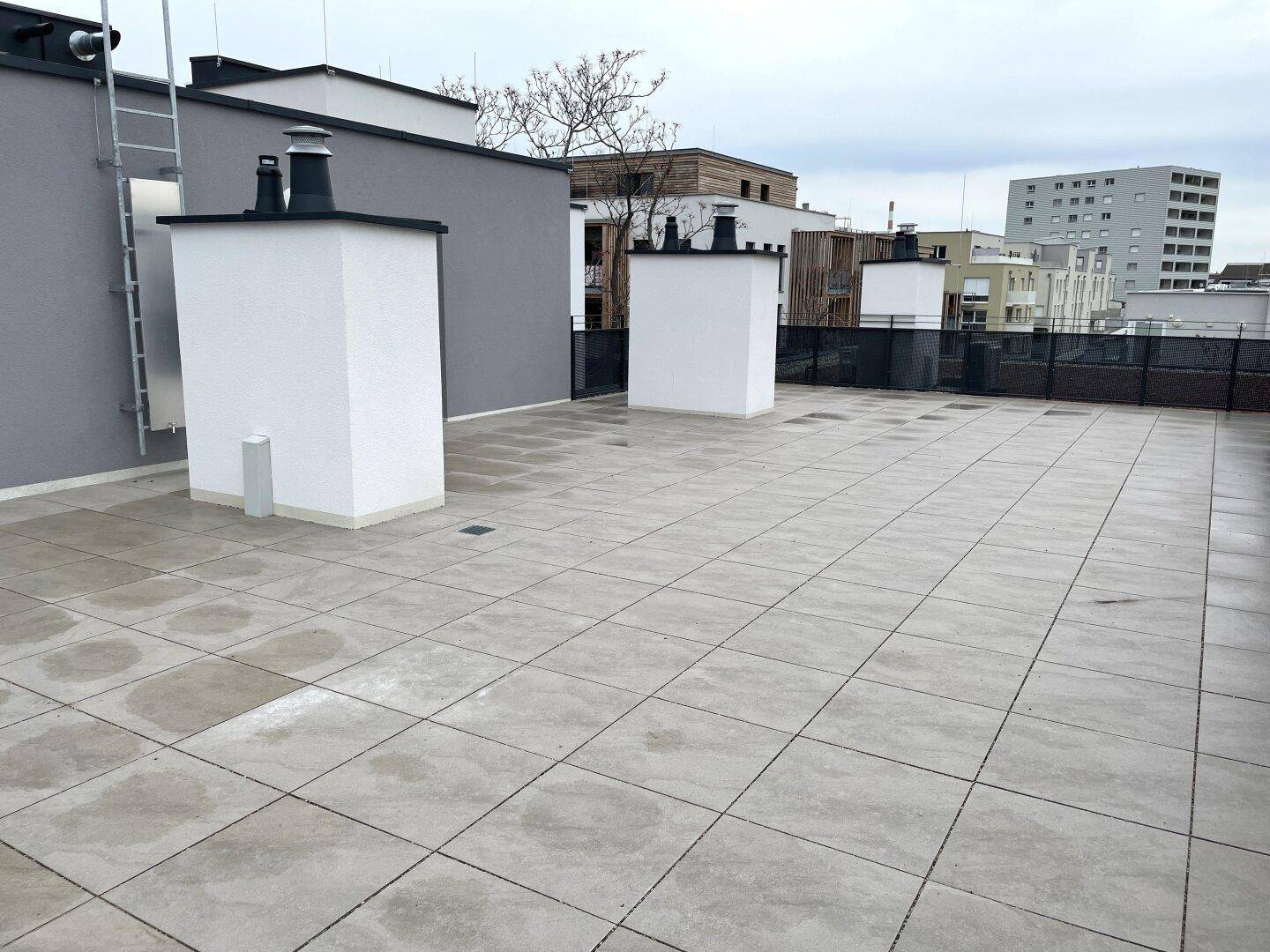 Geräumige Dachterrasse mit grauen Fliesen und weißen Aufbauten unter bewölktem Himmel.