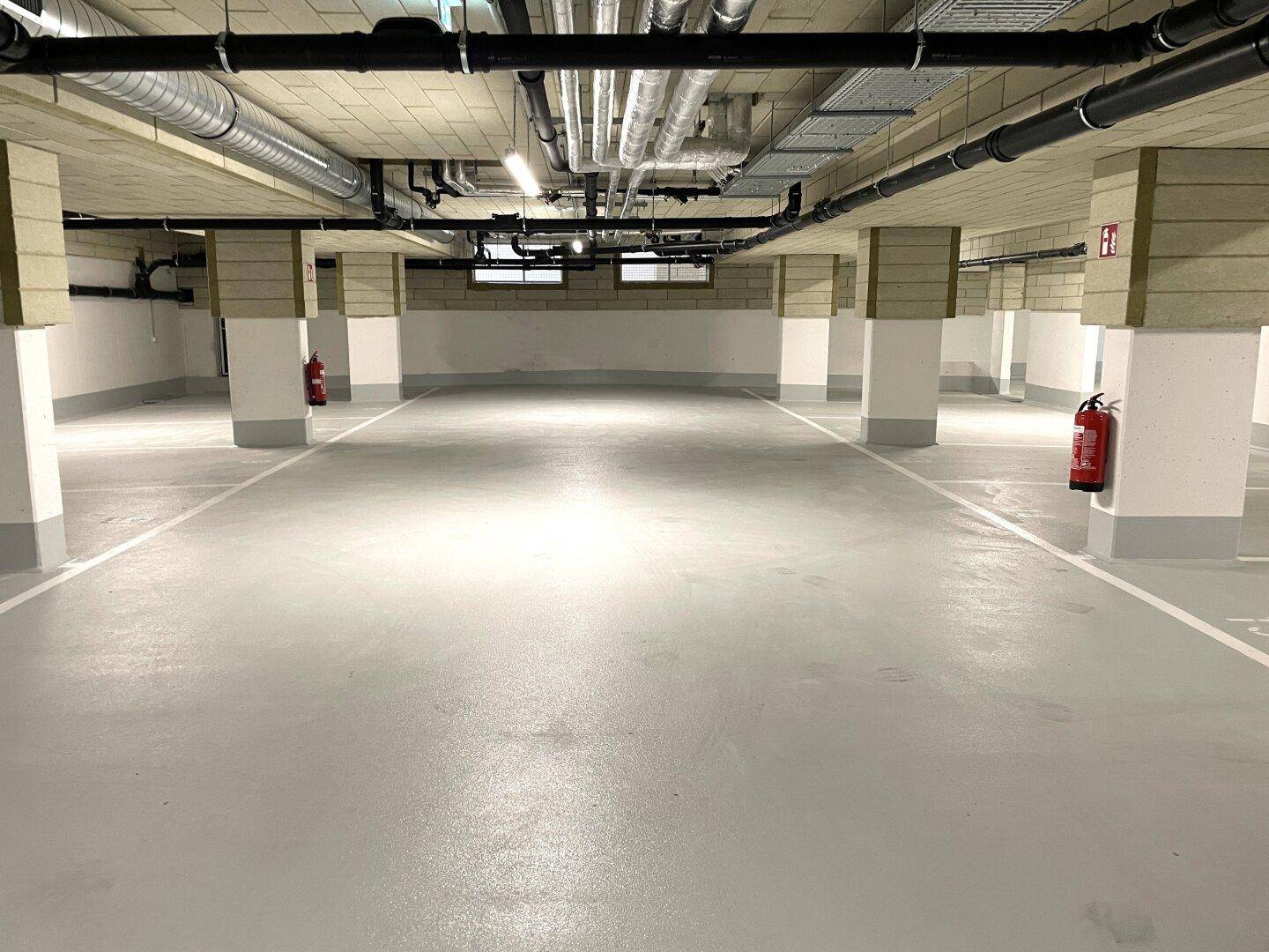 Weitläufige Tiefgarage mit vielen Parkplätzen, Säulen und sichtbaren Versorgungsleitungen an der Decke.