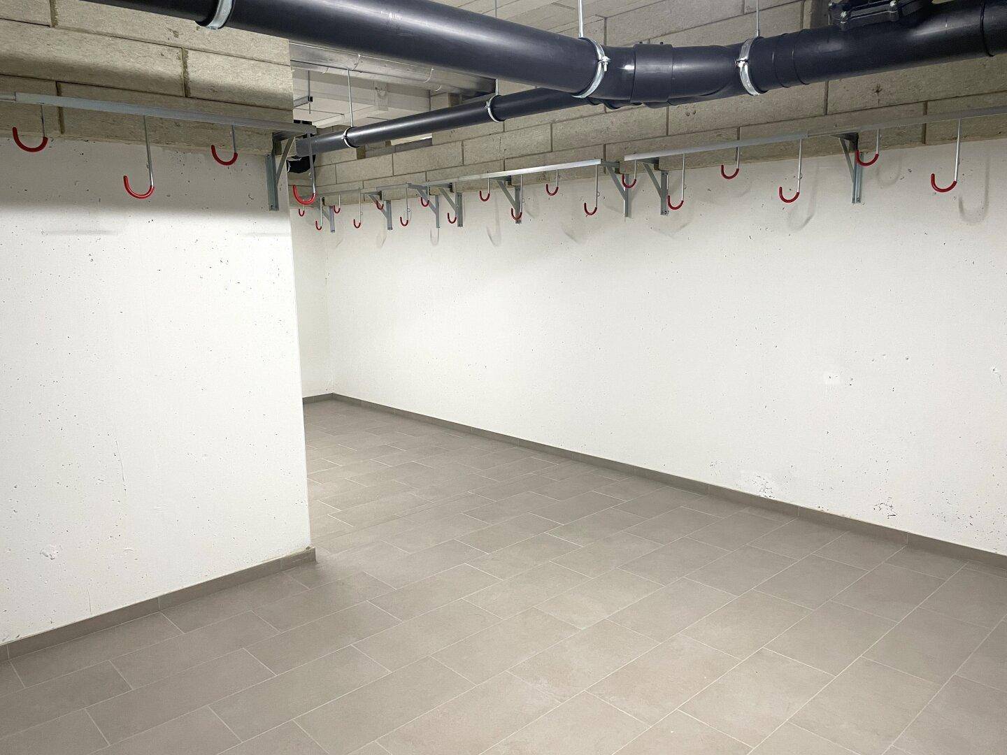 Ein Fahrradraum mit Fliesenboden, Haken an der Wand und sichtbaren Rohren an der Decke.