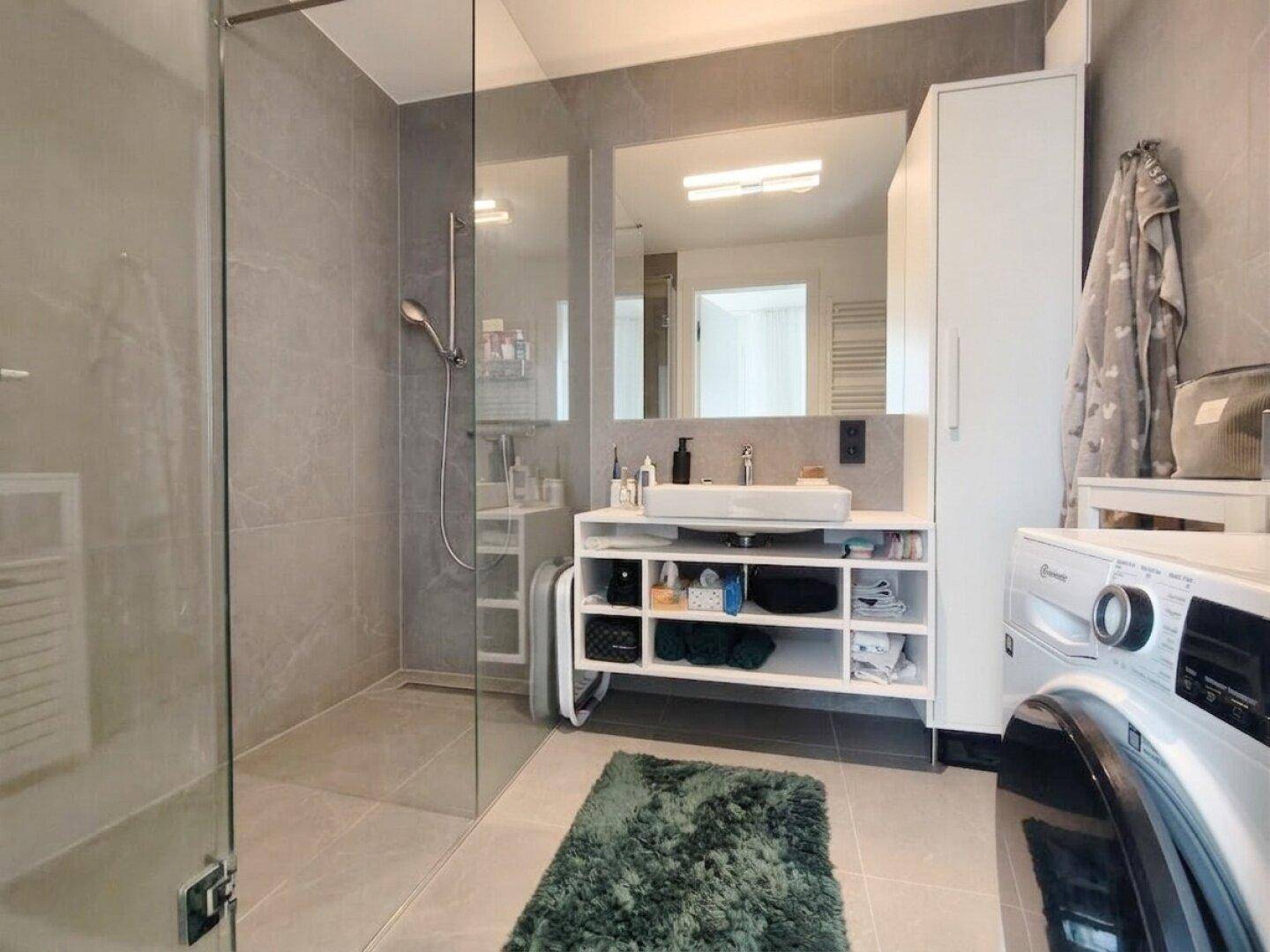 Modernes Badezimmer mit großformatigen grauen Fliesen, begehbarer Dusche und Waschmaschine.