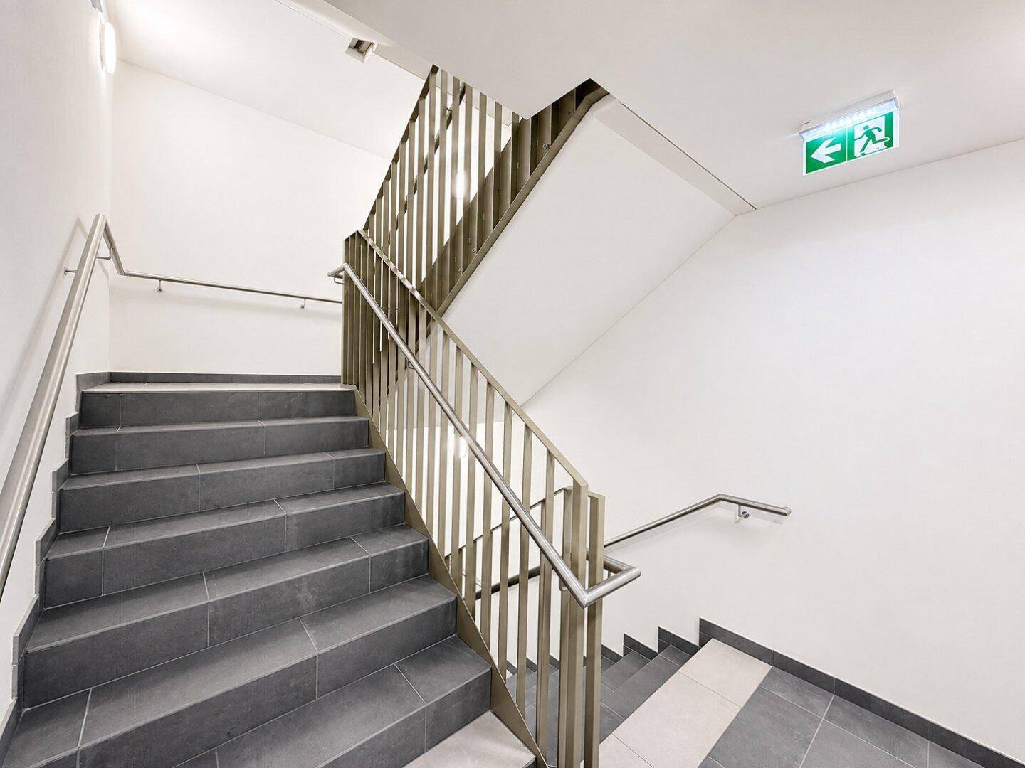 Modernes Treppenhaus mit grauen Fliesen, Edelstahlgeländer und hohen Decken in einem Neubau.
