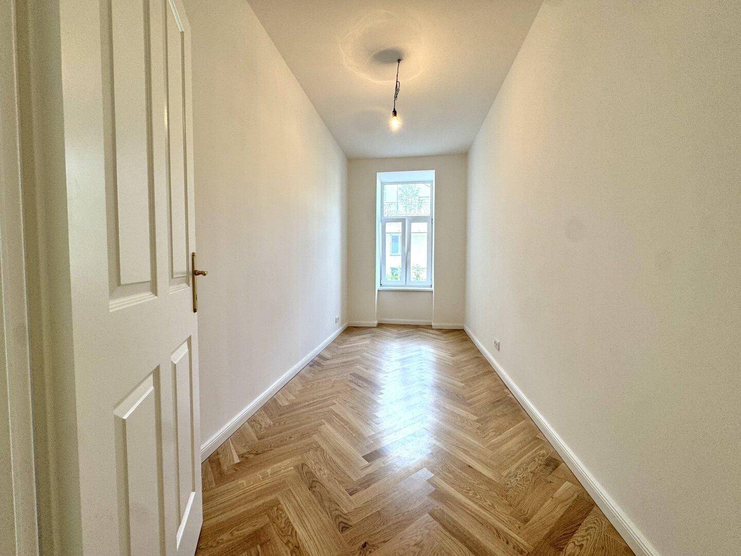 Schmaler, heller Raum mit Fischgrätparkett und einem Fenster, geeignet als Schlafzimmer oder Arbeitszimmer.