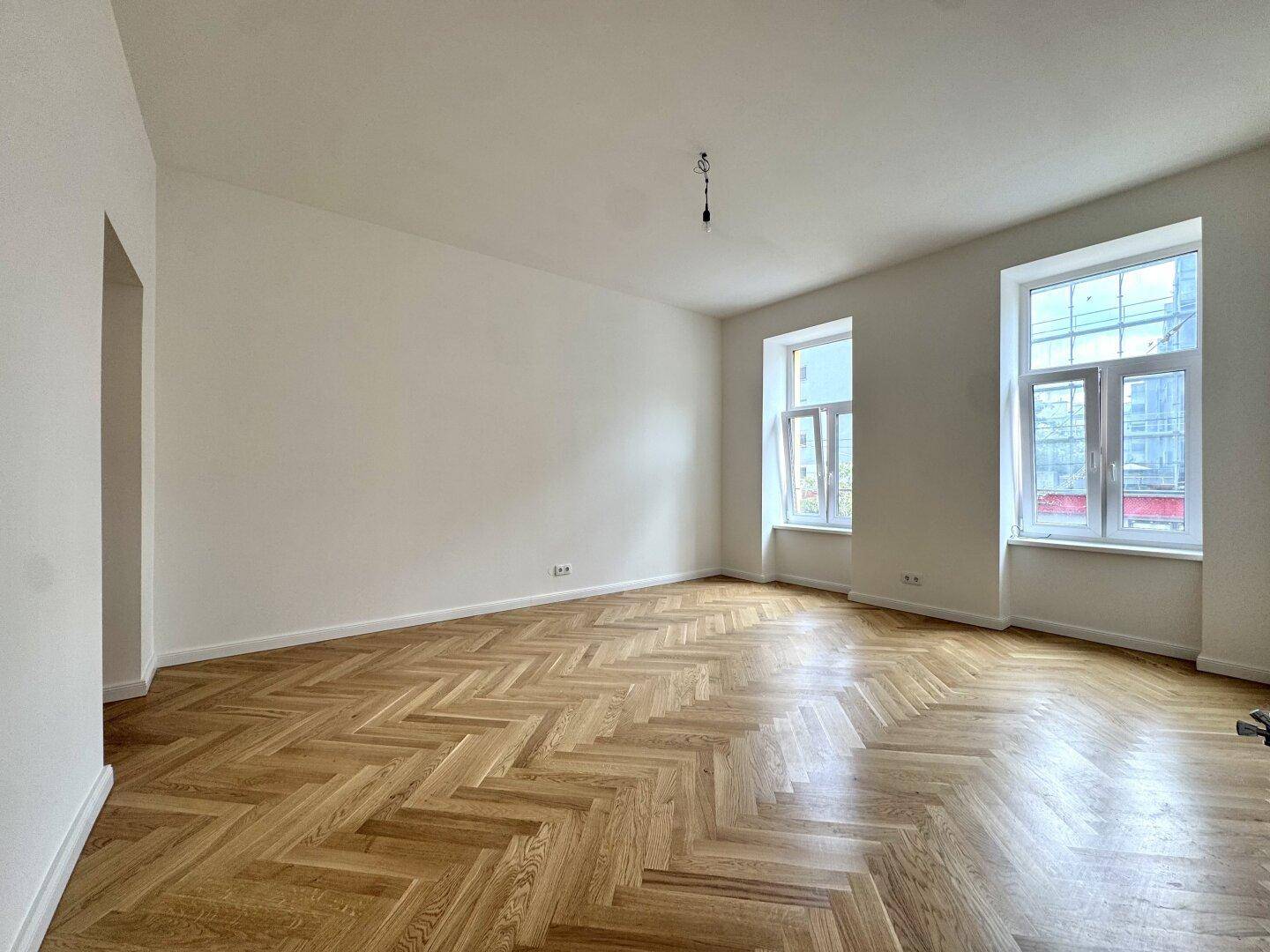 Geräumiges, helles Zimmer mit klassischem Fischgrätparkett und großen Fenstern, ideal für Wohnzwecke.