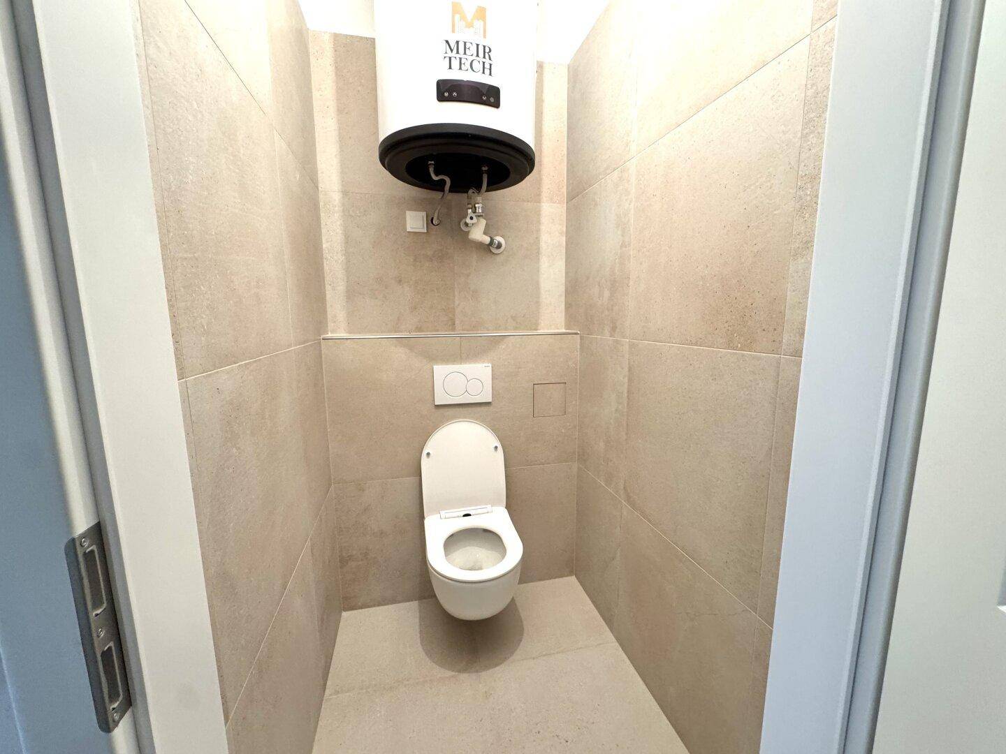 Separates WC mit Wand-WC, beigefarbenen Fliesen und Warmwasserboiler.