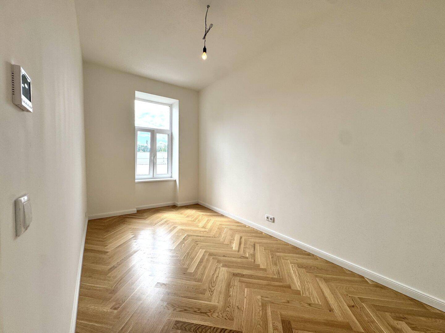 Freundliches Zimmer mit Fischgrätparkett und einem großen Fenster, bietet viel Tageslicht.