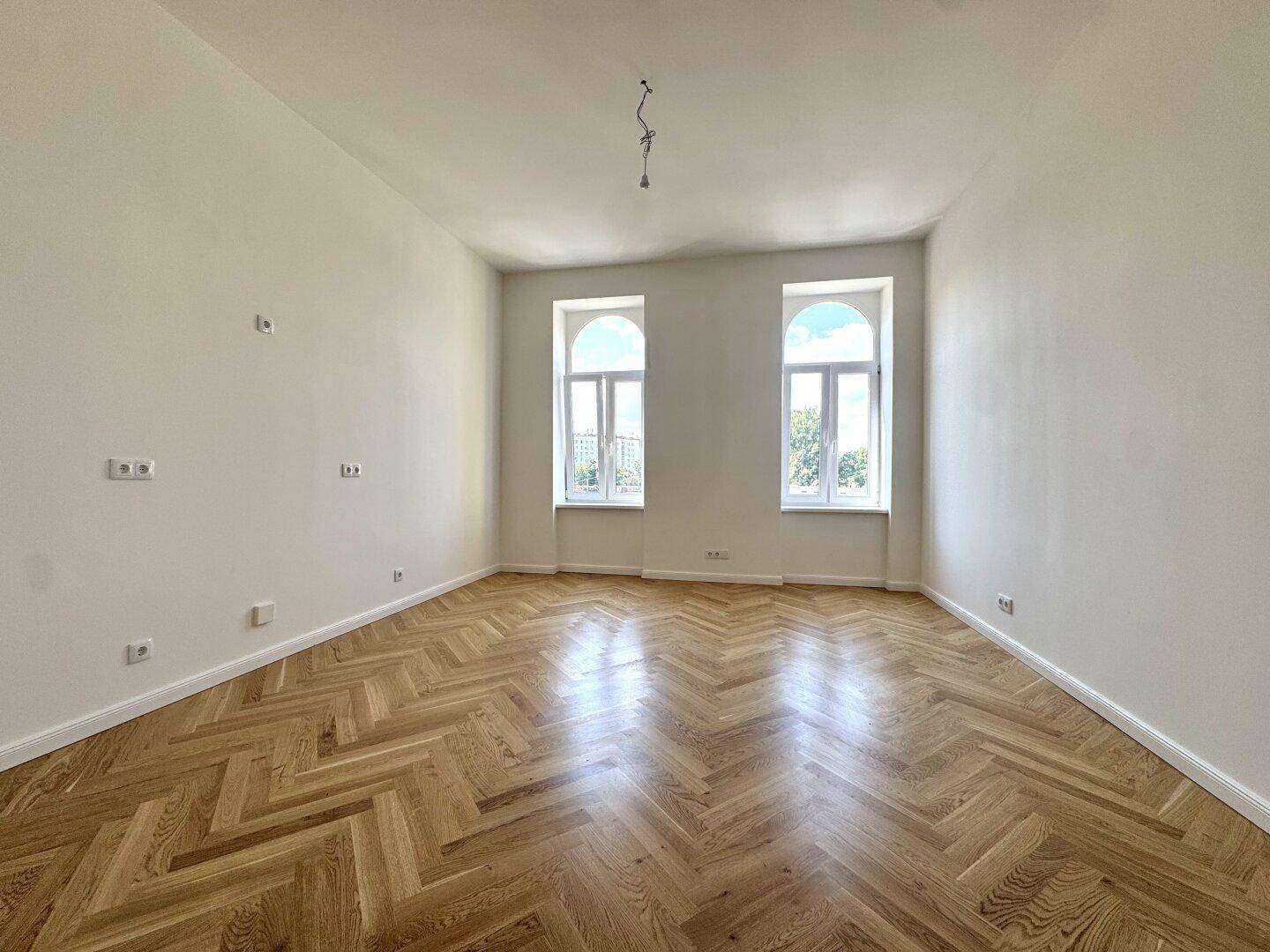 Geräumiges, unmöbliertes Zimmer mit Fischgrätparkett und zwei hohen Bogenfenstern.