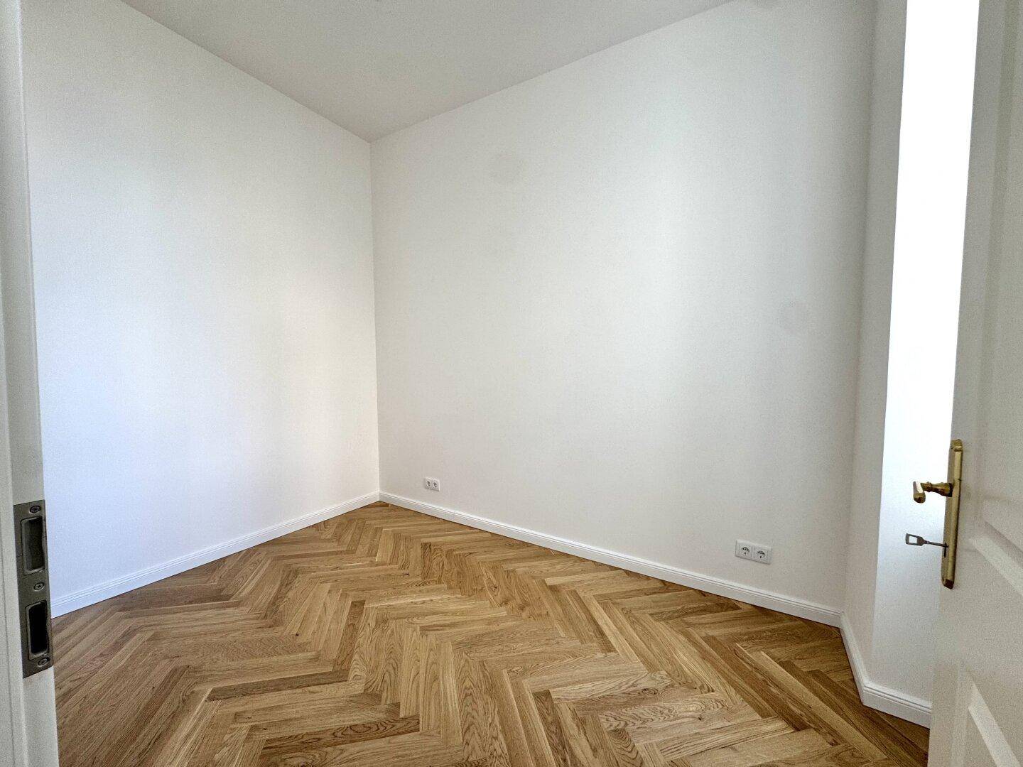 Kleineres, unmöbliertes Zimmer mit Fischgrätparkett und weißen Wänden.