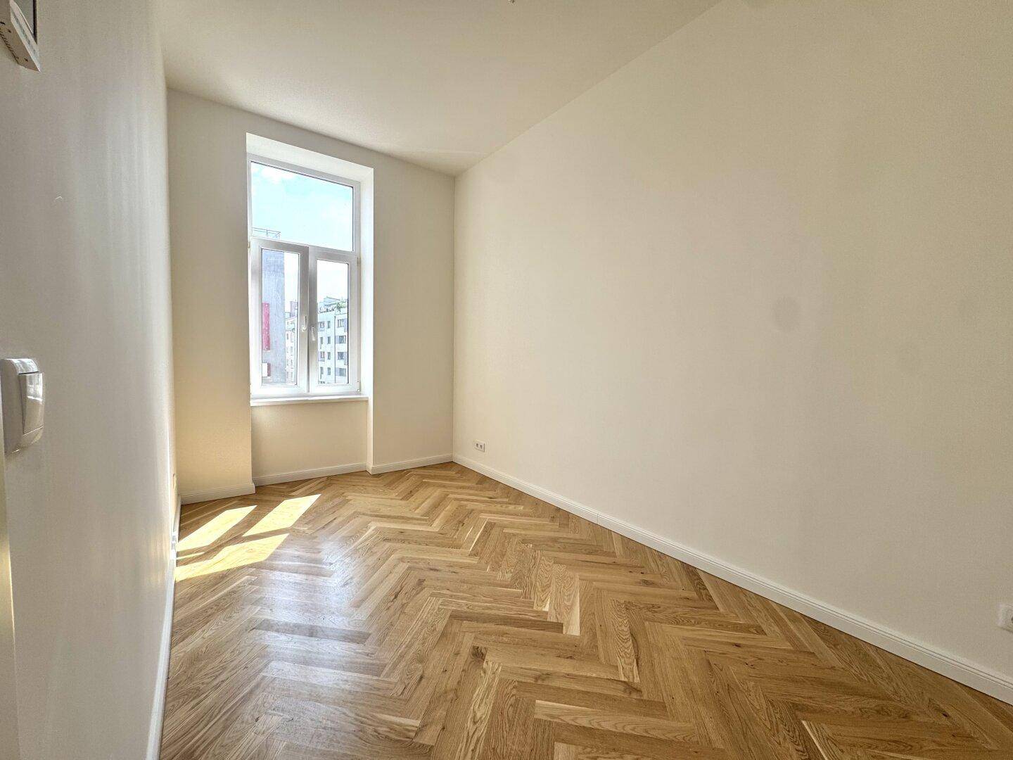 Lichtdurchflutetes Zimmer mit elegantem Fischgrätparkett und einem hohen Fenster, ideal als Schlaf- oder Arbeitszimmer.