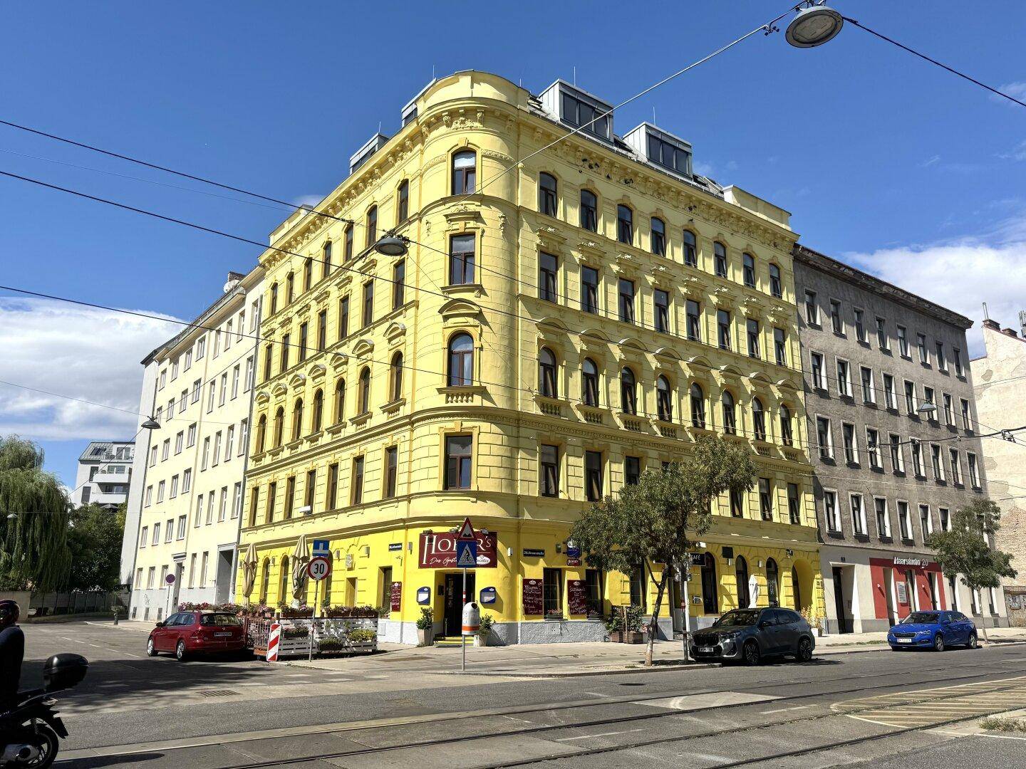 Historisches Eckgebäude mit gelber Fassade und Geschäften im Erdgeschoss an einer belebten Straße.