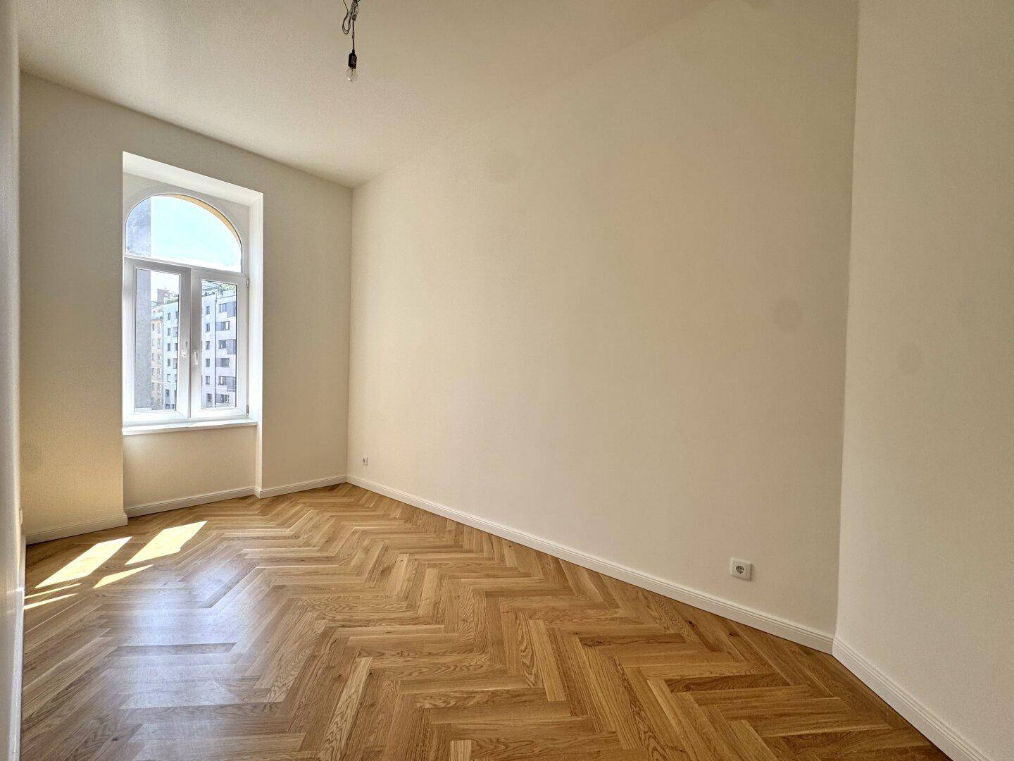 Gemütliches Zimmer mit Fischgrätparkett und einem großen Bogenfenster, ideal als Schlafzimmer.