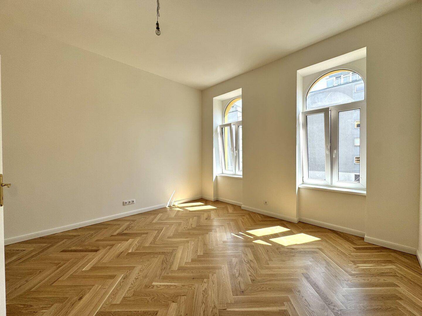 Helles Zimmer mit klassischem Fischgrätparkett und zwei hohen Bogenfenstern.