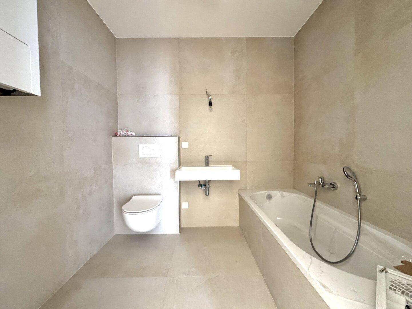Modernes Badezimmer mit beigefarbenen Fliesen, wandhängendem WC, Einzelwaschbecken und Badewanne.