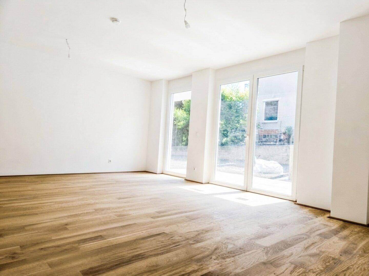 Heller, leerer Wohnraum mit großen Fenstern, Holzboden und Zugang zum Außenbereich.