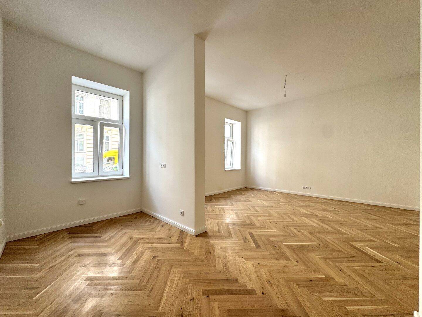 Geräumiges Zimmer mit klassischem Fischgrätparkett und viel Tageslicht durch große Fenster.