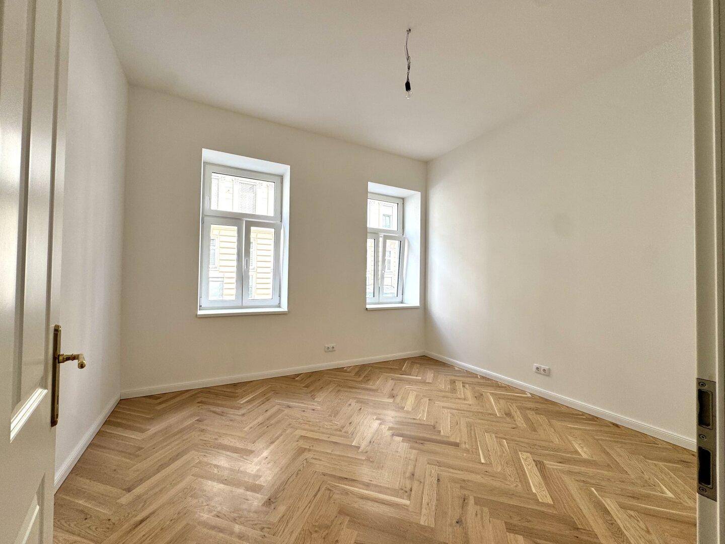 Ruhiges Schlafzimmer mit hellem Fischgrätparkett und zwei Fenstern für angenehmes Tageslicht.